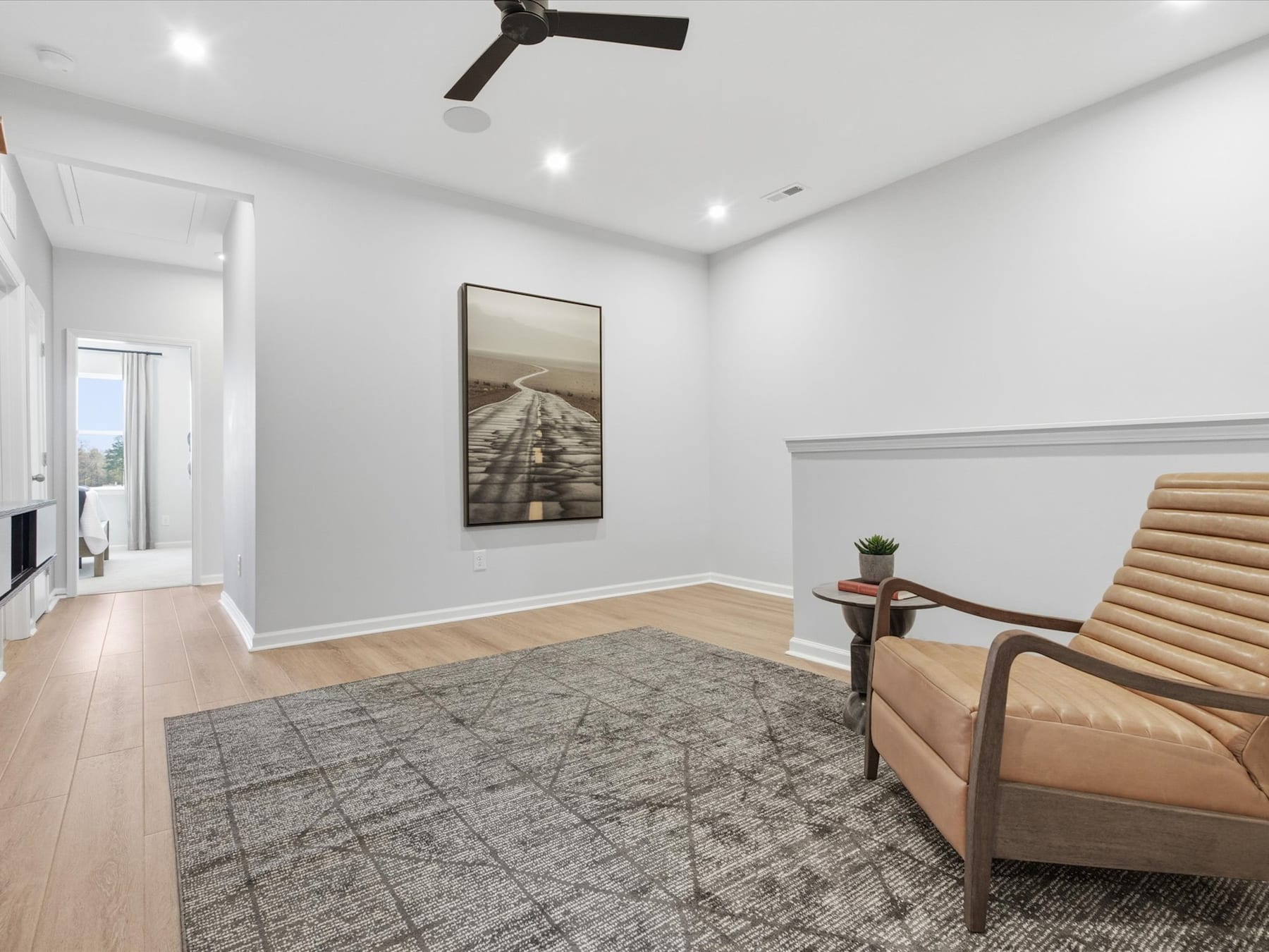 A spacious and well-lit room with a wooden floor, a ceiling fan, and a framed artwork on the wall. In the foreground, there is a comfortable-looking armchair.