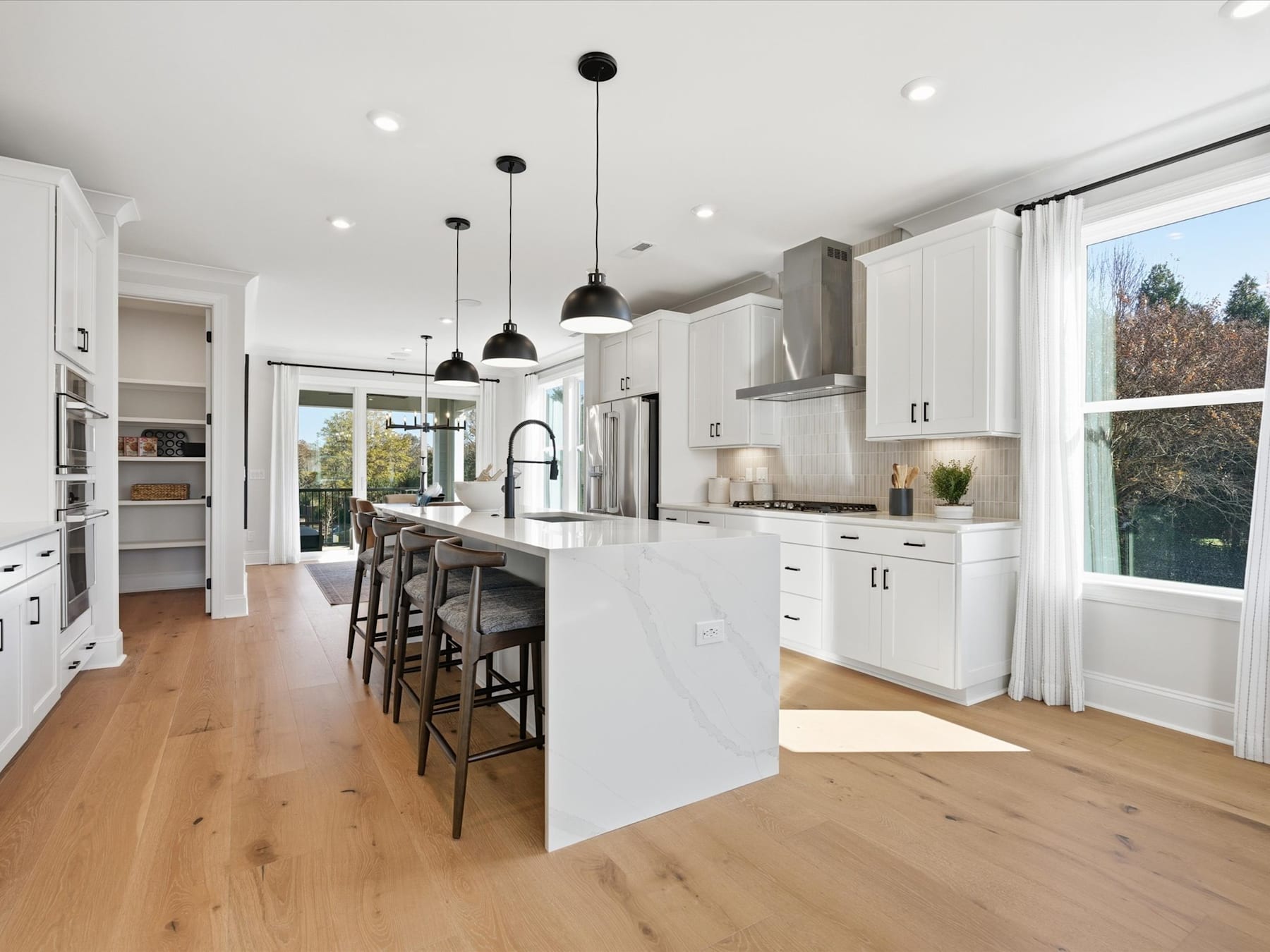 A modern, open-concept kitchen with white cabinets, a large island, and pendant lighting, set against a backdrop of large windows overlooking a scenic outdoor view.