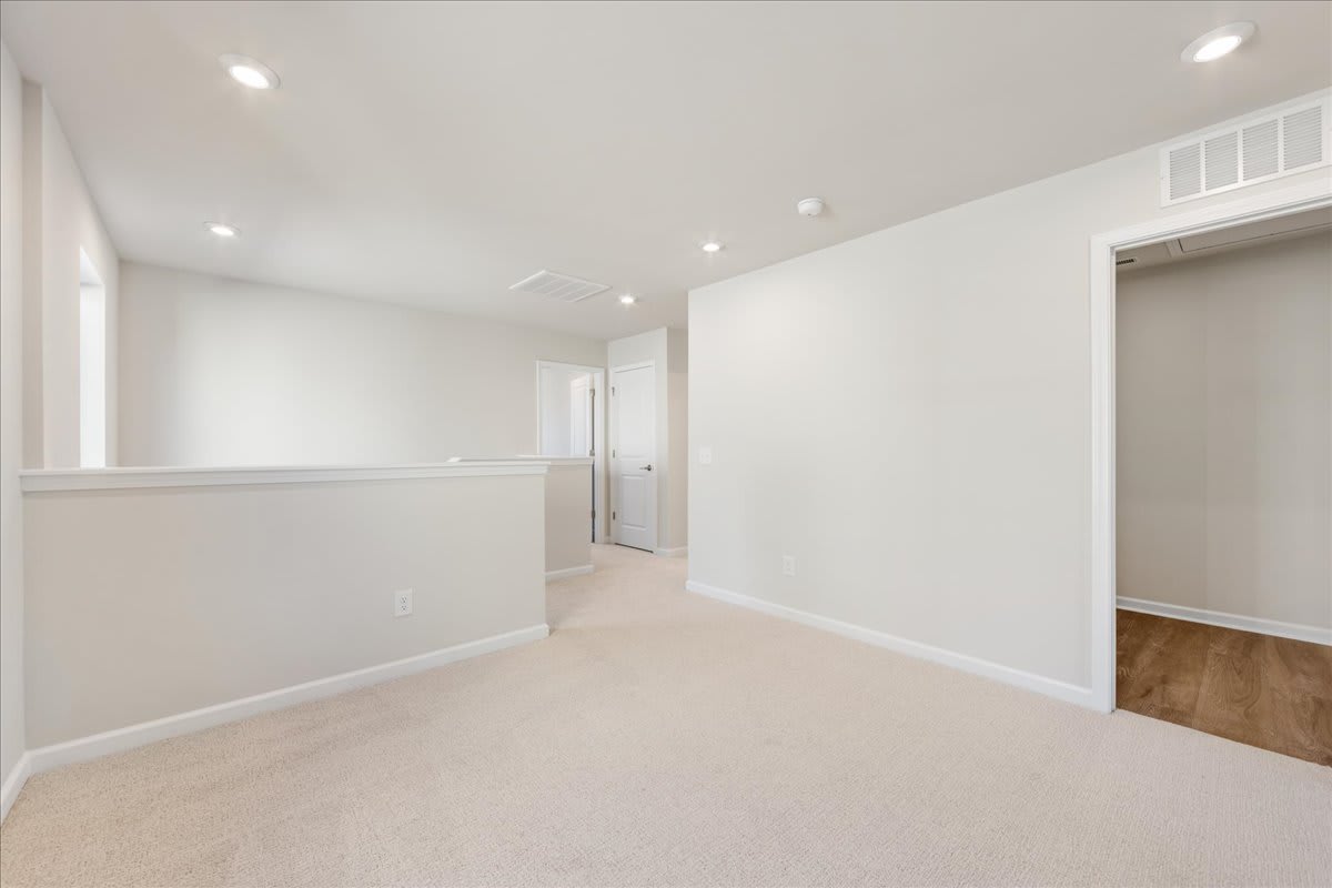 A bright, spacious room with white walls, recessed lighting, and a carpeted floor. The room appears to be a hallway or entryway, with a doorway visible in the background.