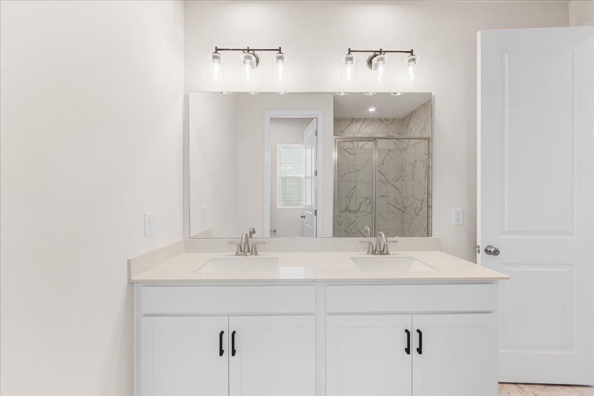 A modern, minimalist bathroom with a double vanity, white cabinets, and a large mirror above it, set against a backdrop of white walls and marble-like tiles.