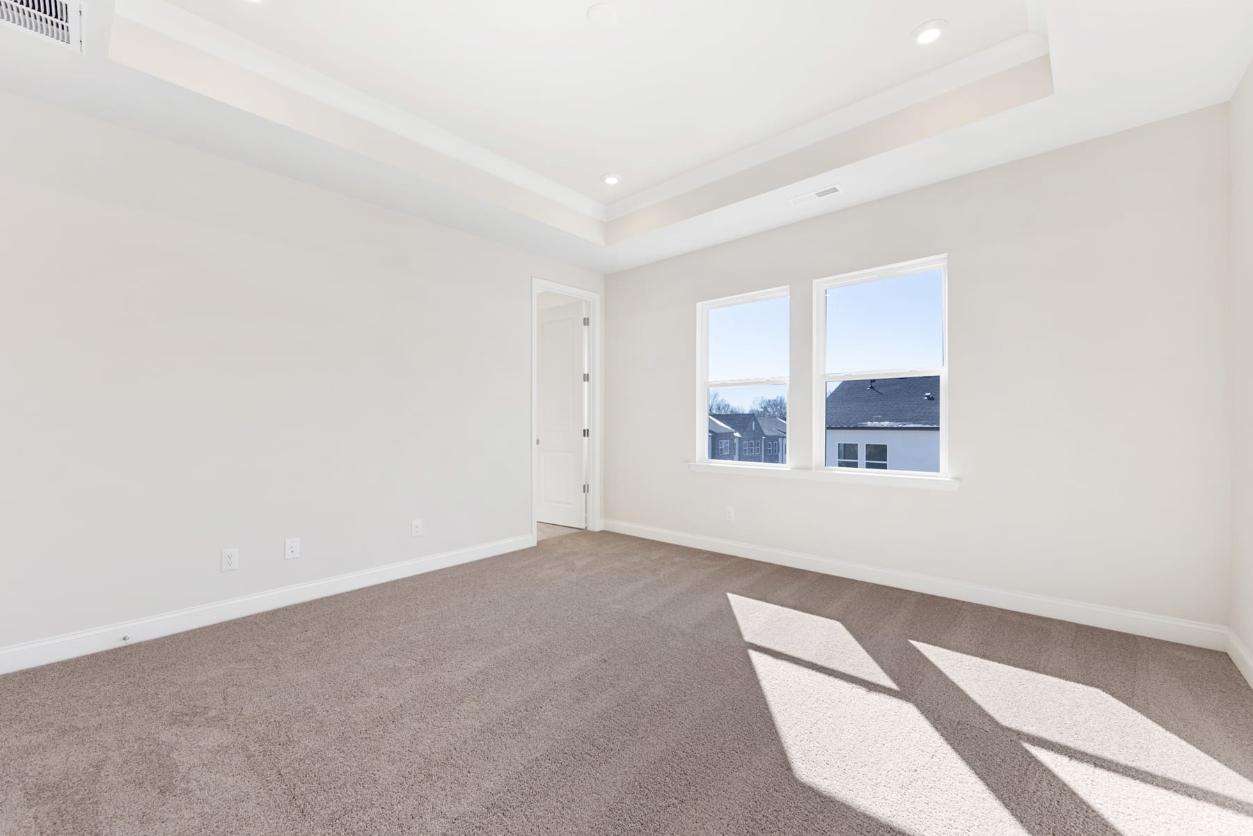 An empty, bright room with white walls, a carpeted floor, and large windows providing natural light.