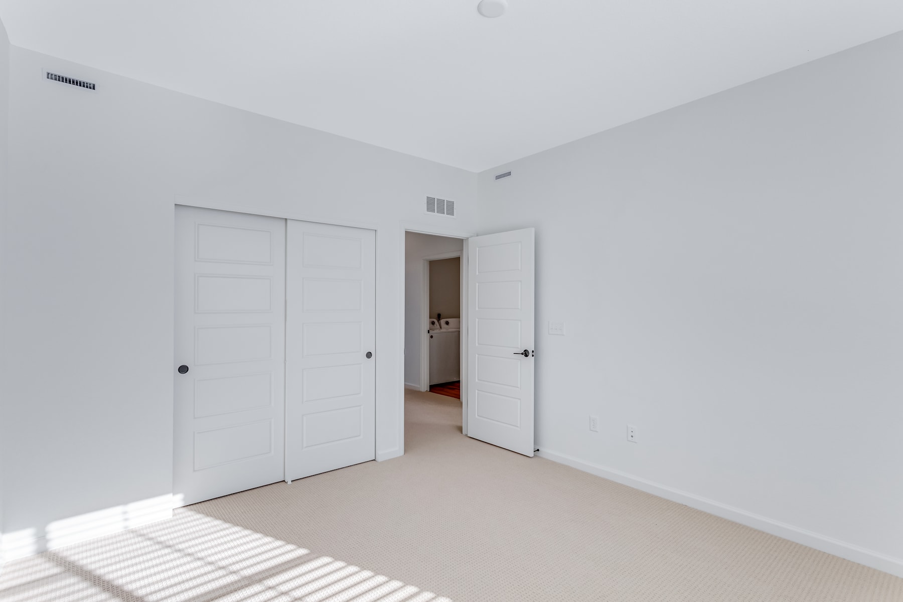 A simple, minimalist room with white walls, a closed door, and a partially open door leading to another room.