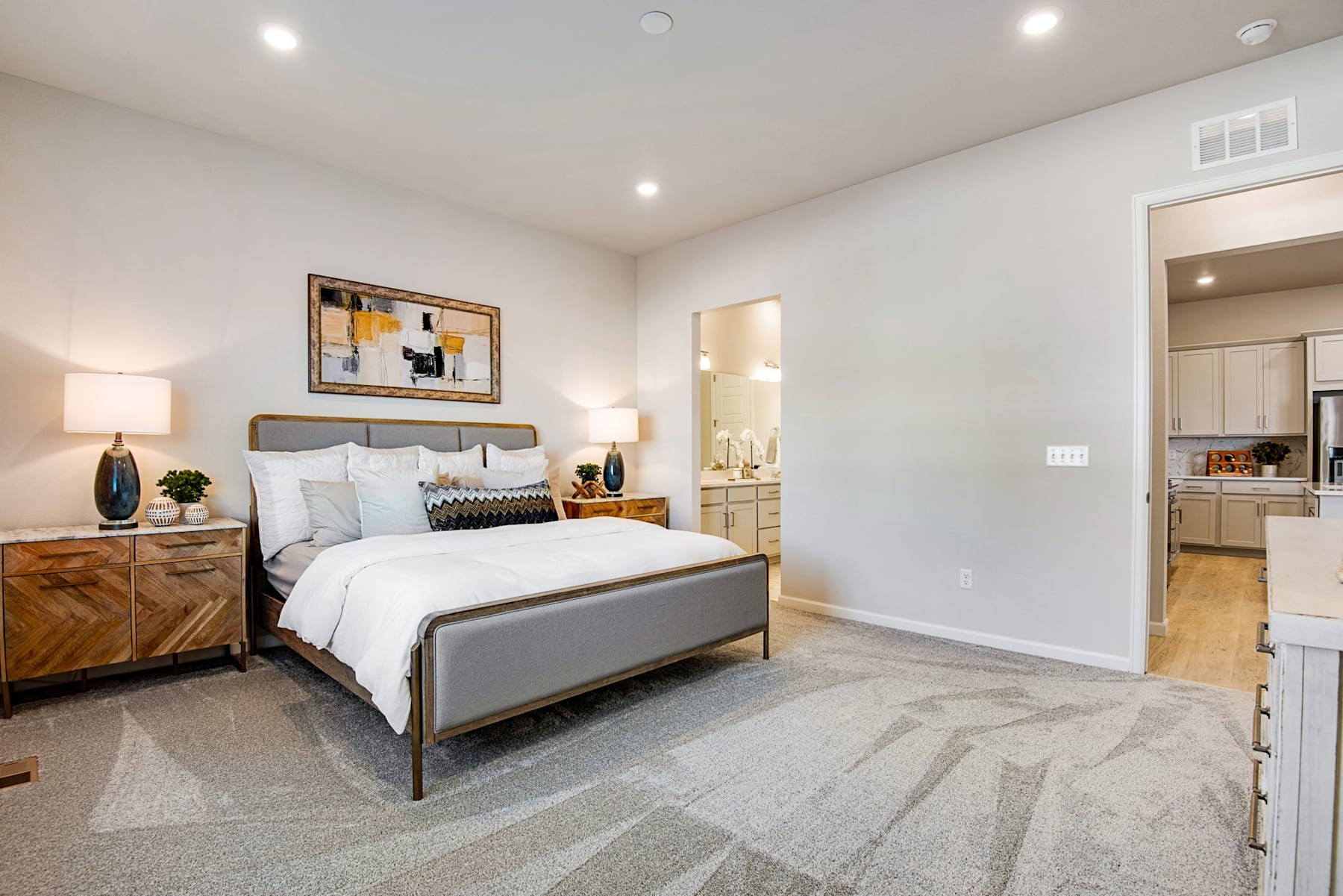 A spacious and well-designed bedroom with a large bed, wooden furniture, and a modern art piece on the wall, leading to an open kitchen area.