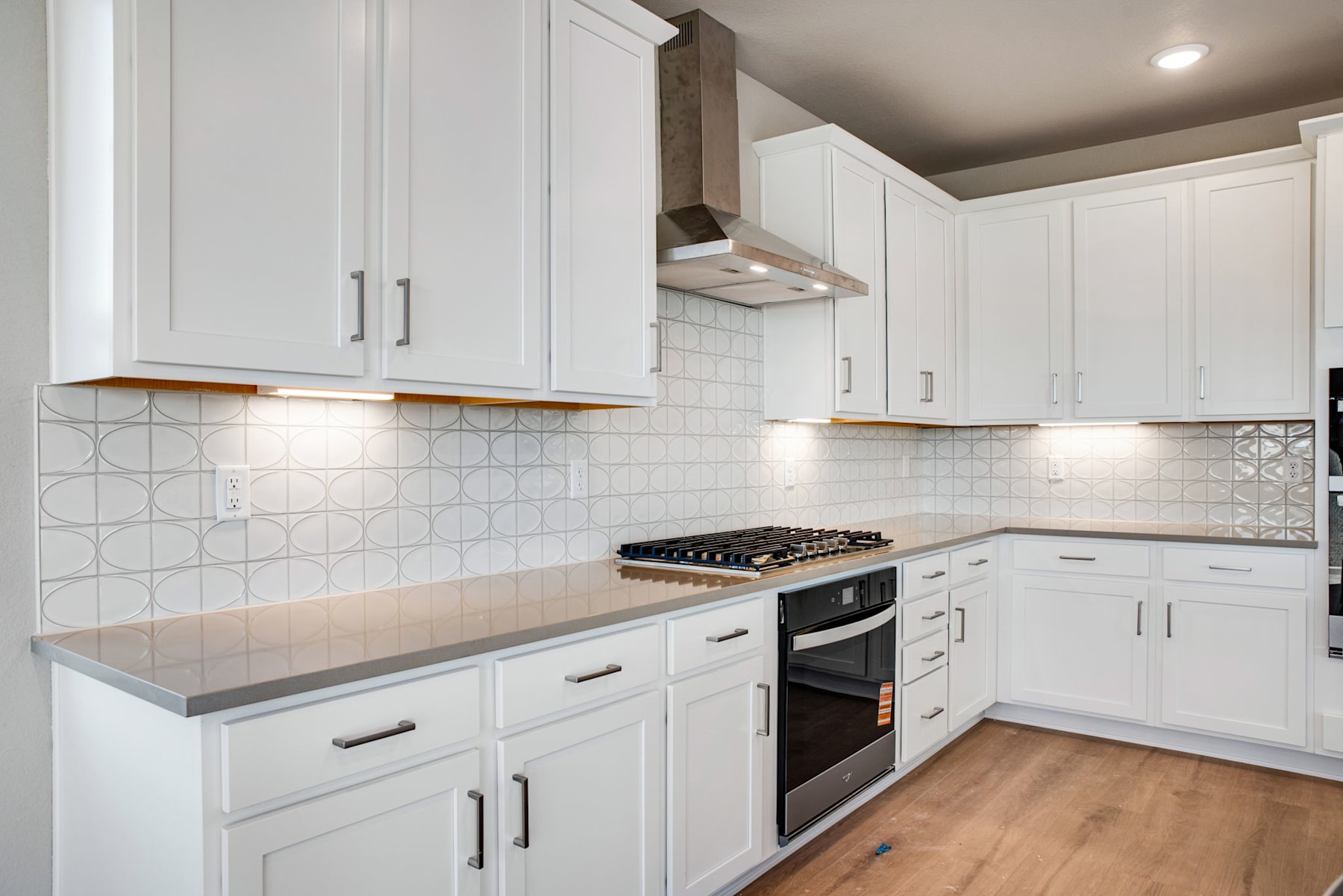 A modern, well-lit kitchen with white cabinets, a gas stove, and a tiled backsplash, set against a hardwood floor.