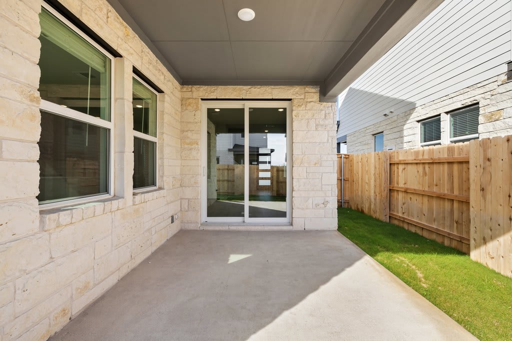 A paved walkway leads to a glass-paneled door, flanked by a wooden fence and surrounded by a grassy yard, with a stone exterior wall visible in the background.