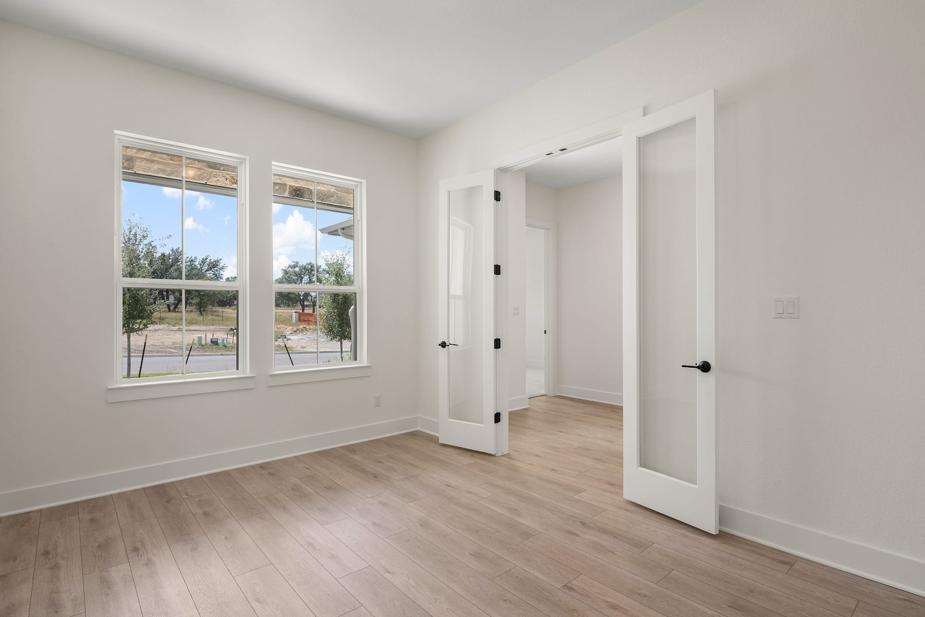A bright and airy room with large windows, white walls, and a wooden floor. The room appears to be a bedroom or living space with a door leading to another area.
