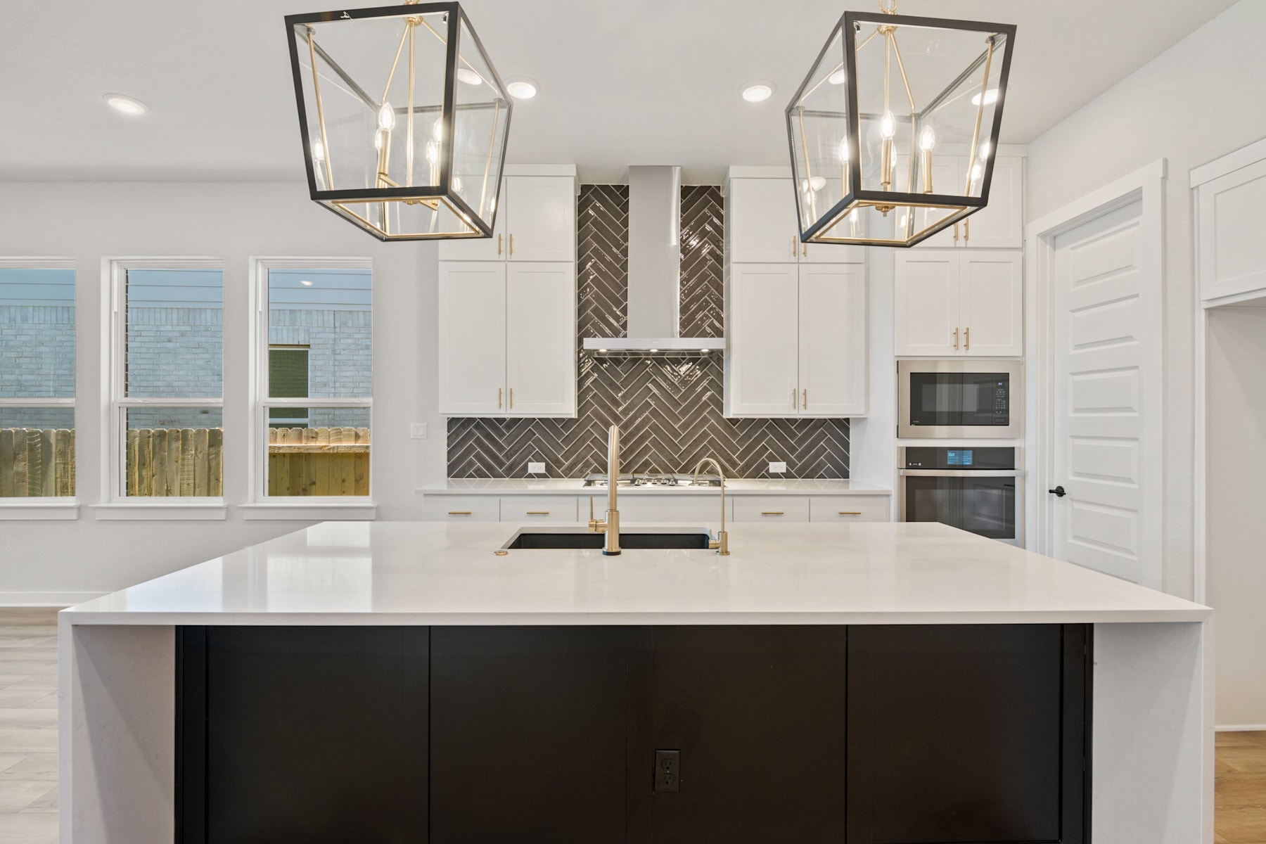 A modern, open-concept kitchen with a large white countertop island, black cabinetry, and two pendant light fixtures with a geometric design.