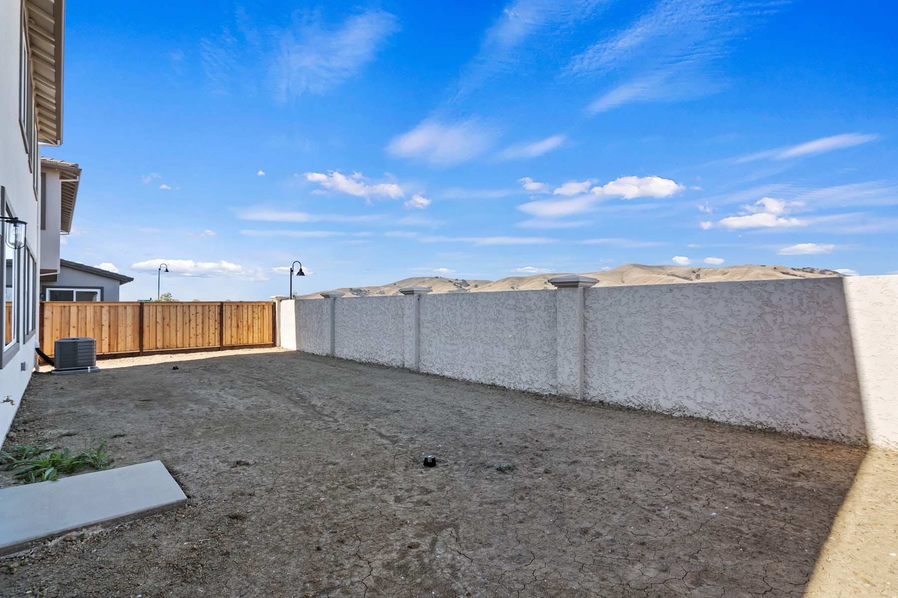 A paved backyard area with a wooden fence, surrounded by a clear blue sky with scattered clouds.