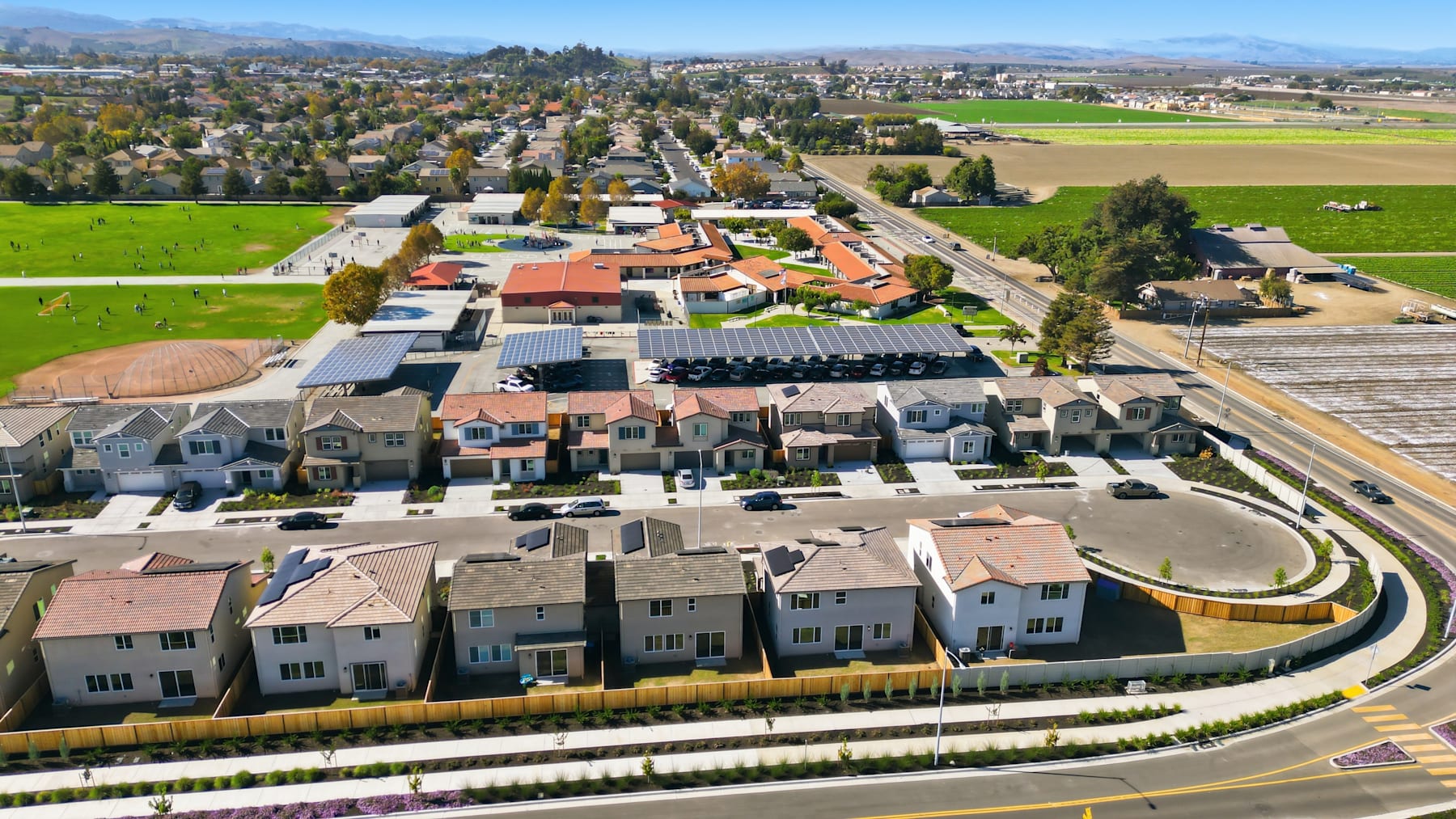 A residential neighborhood with rows of houses and townhouses, surrounded by lush greenery, rolling hills, and a clear blue sky in the background.