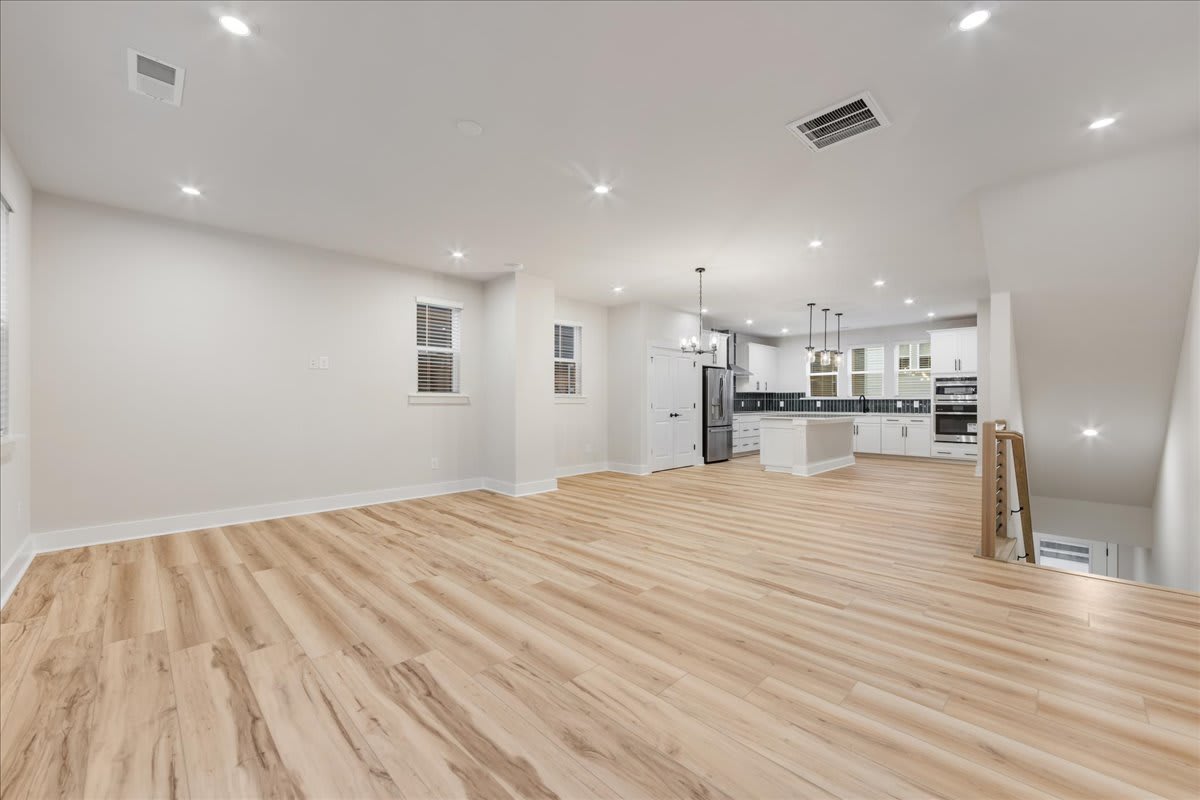 A spacious, open-concept living area with a modern kitchen visible in the background, featuring hardwood floors, recessed lighting, and a clean, minimalist design.