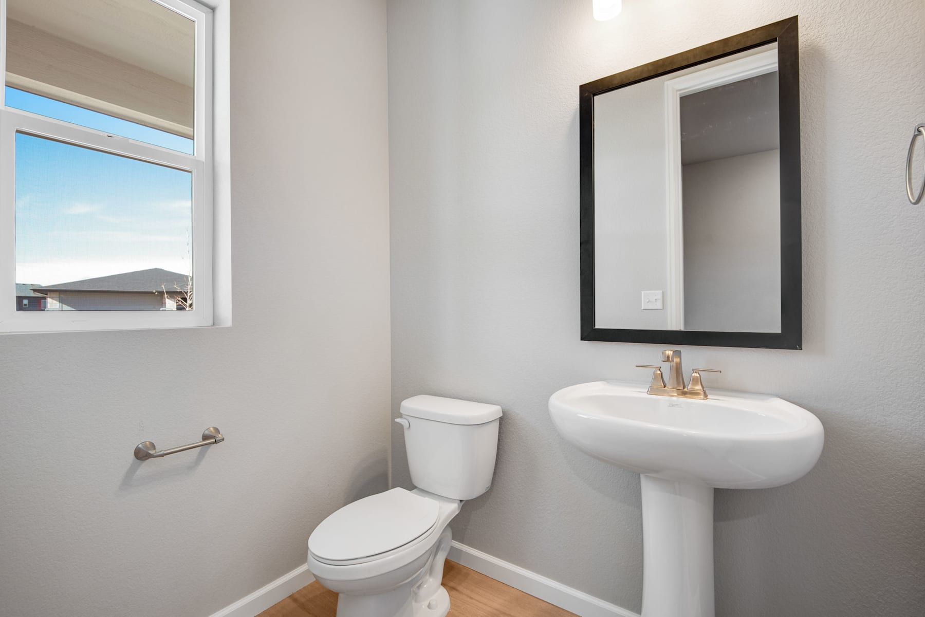 A modern and minimalist bathroom with a white pedestal sink, a toilet, and a framed mirror on the wall. The room has a large window that provides natural light and a view of the exterior.