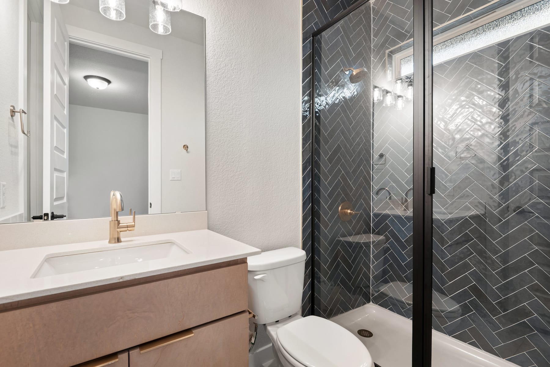 A modern bathroom with a white vanity, a toilet, and a glass-enclosed shower with gray herringbone tile walls.
