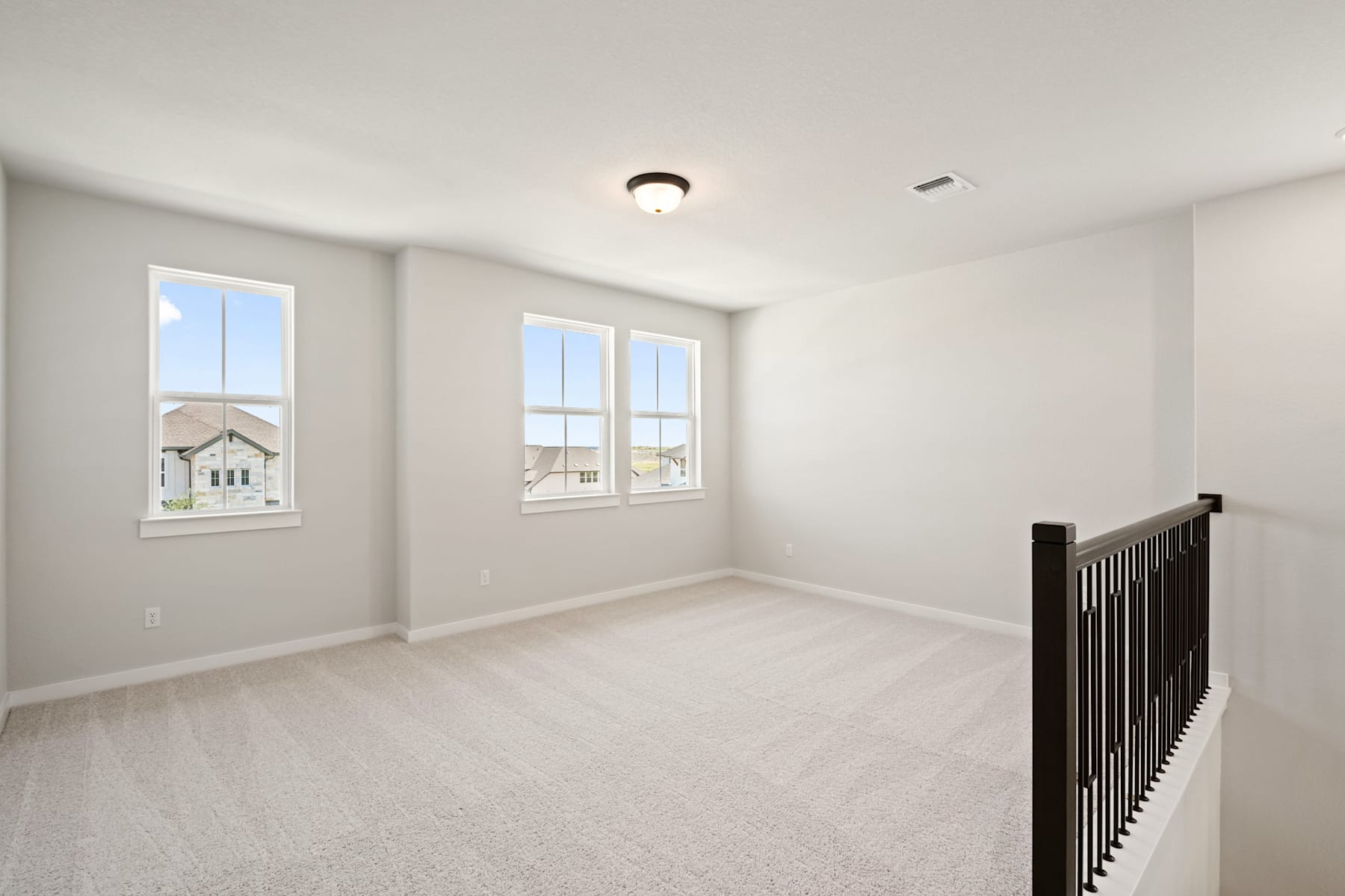 A bright, empty room with white walls, a ceiling light fixture, and a wooden staircase railing in the foreground.