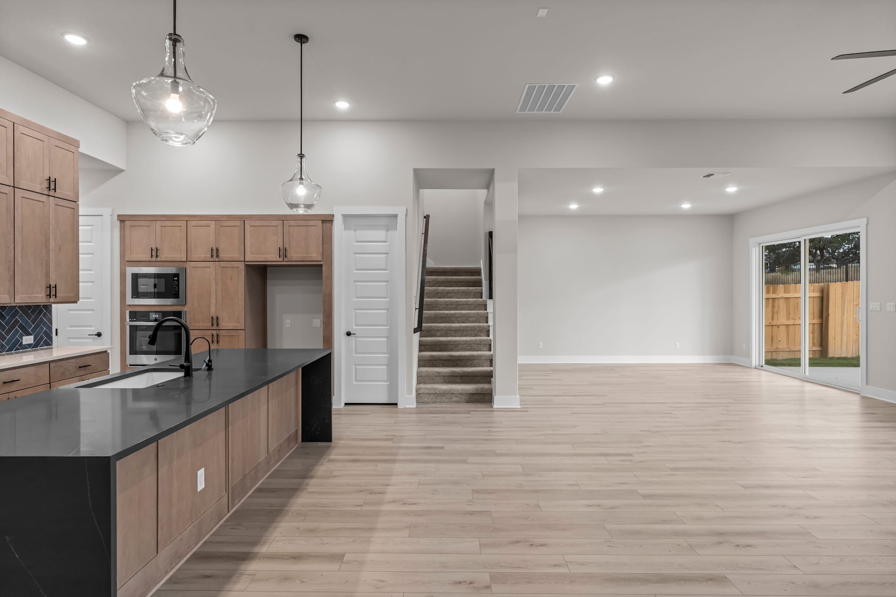A modern, open-concept kitchen and living space with light wood cabinets, black countertops, and pendant lighting fixtures, leading to a staircase and a bright, airy atmosphere.