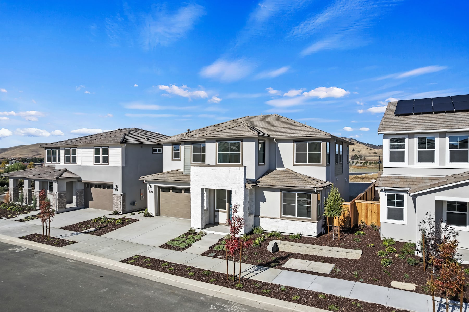 A residential neighborhood with modern, two-story houses featuring gray and white exteriors, landscaped yards, and a clear blue sky with wispy clouds overhead.