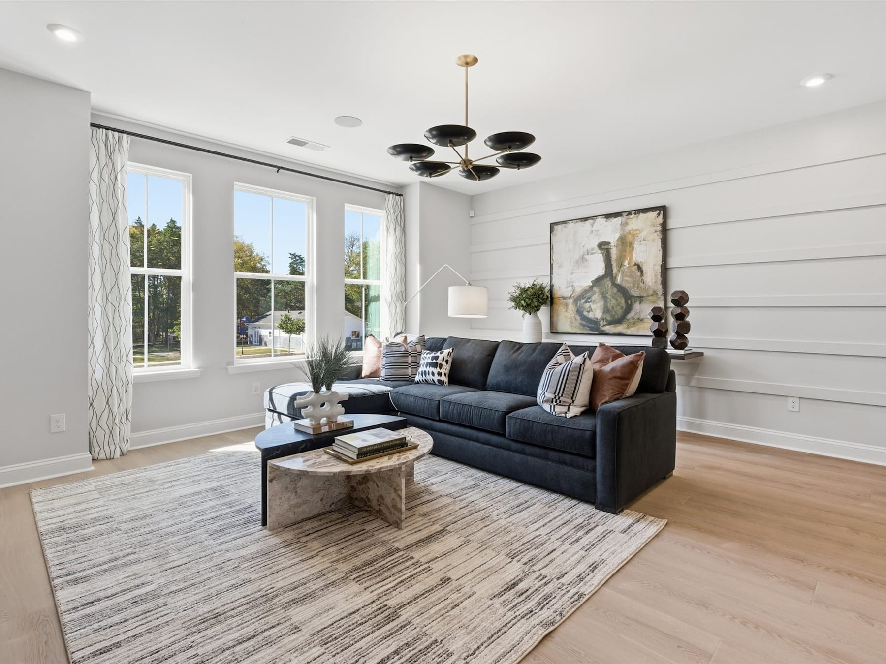 A cozy and modern living room with a large gray sectional sofa, a wooden coffee table, and a decorative chandelier hanging from the ceiling. The room features large windows that allow natural light to flood the space, and the walls are adorned with artwork and other decorative elements.
