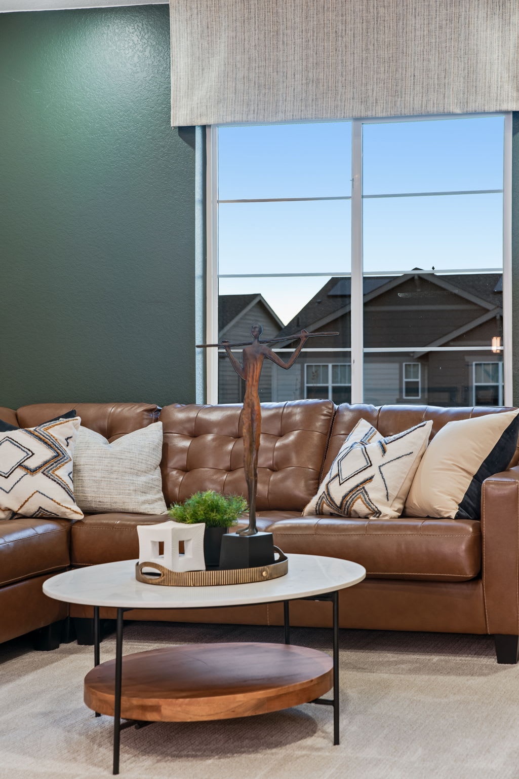 A cozy living room with a large brown leather sectional sofa, a round wooden coffee table, and a view of the exterior buildings through the large windows.