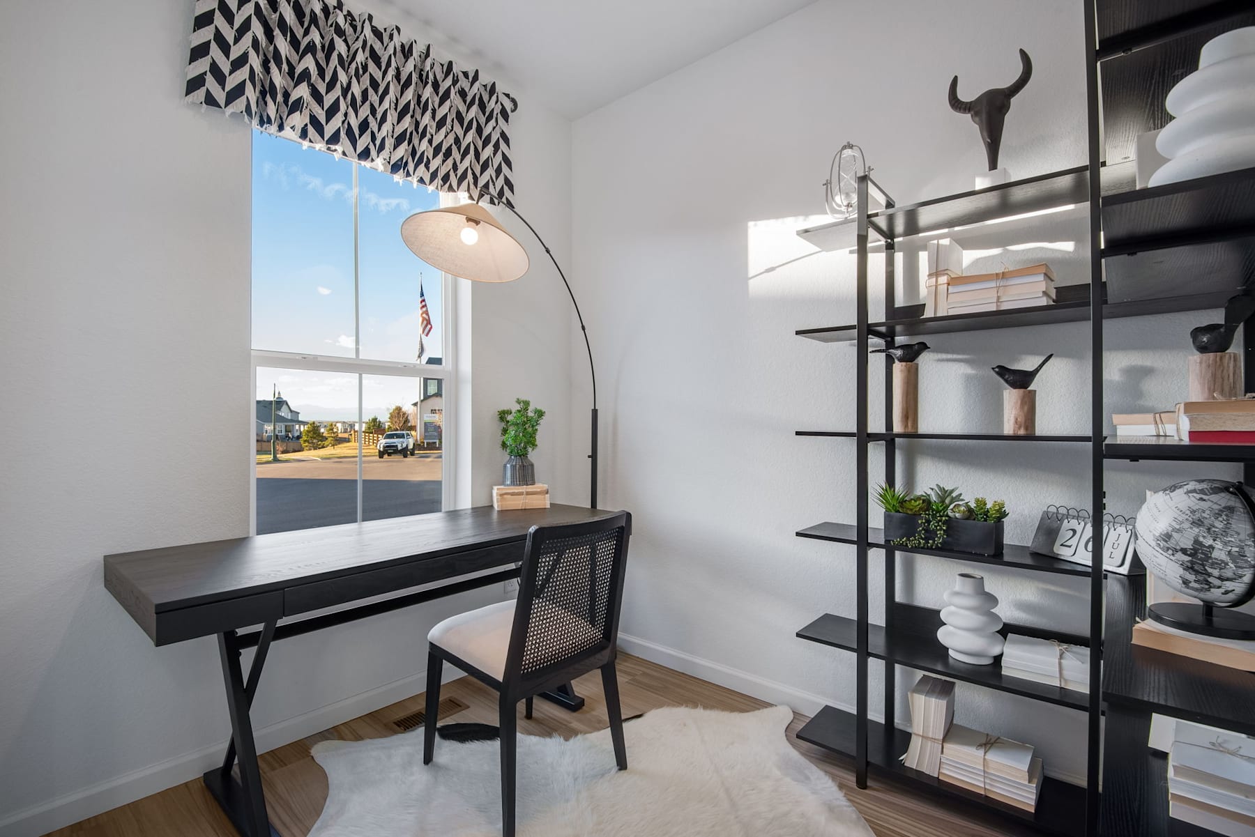 A modern and minimalist home office space with a black desk, a floor lamp, and a shelving unit displaying various decorative items against a backdrop of a large window overlooking an outdoor scene.