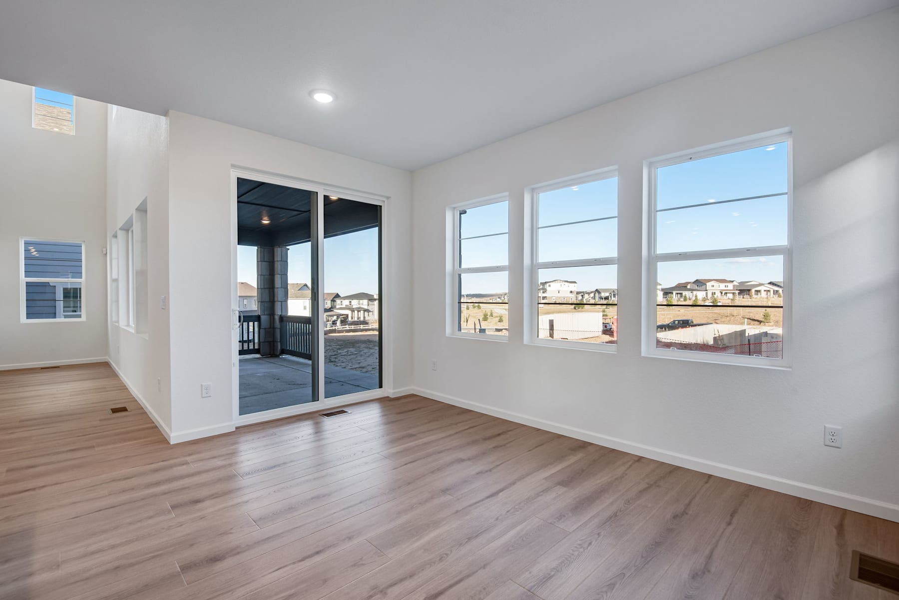 A bright and spacious room with large windows overlooking a residential neighborhood, featuring hardwood floors and a sliding glass door leading to a balcony or patio.