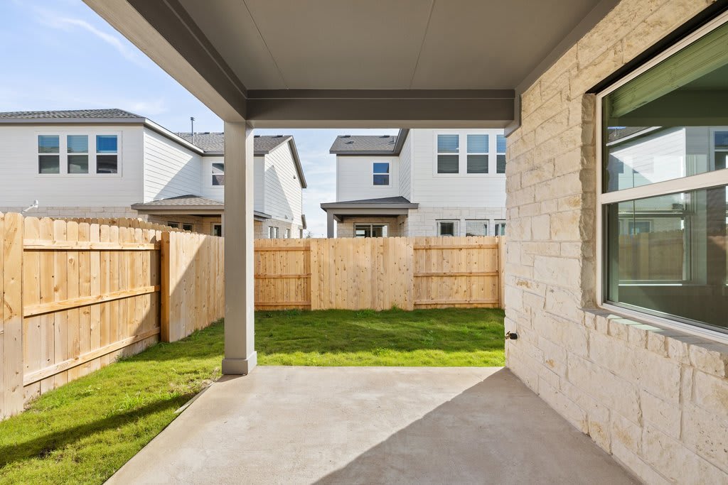 A fenced backyard with a grassy lawn, surrounded by a brick building with windows and a covered patio area.