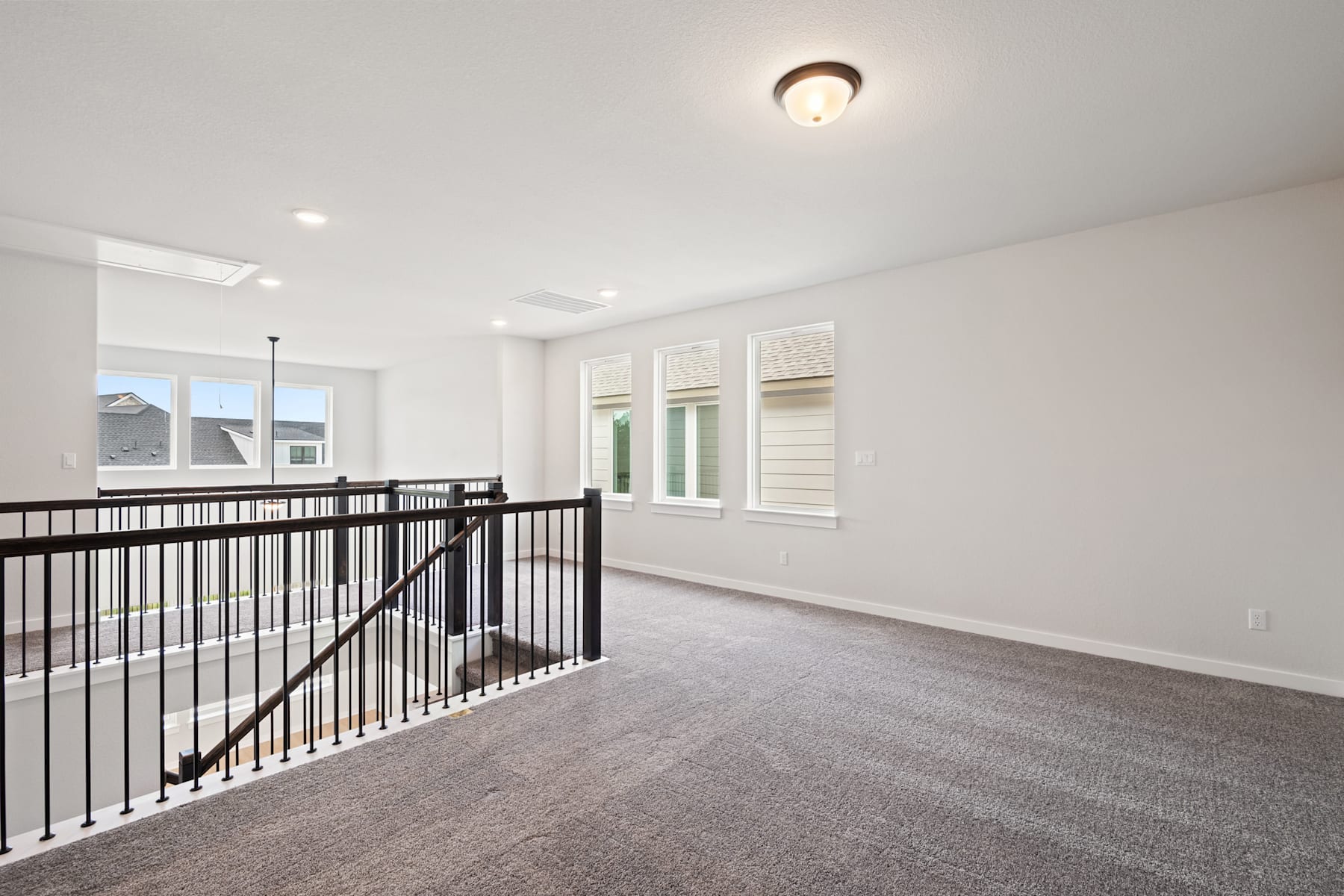 An empty room with a carpeted floor, a railing, and a ceiling light fixture, with windows providing natural light.