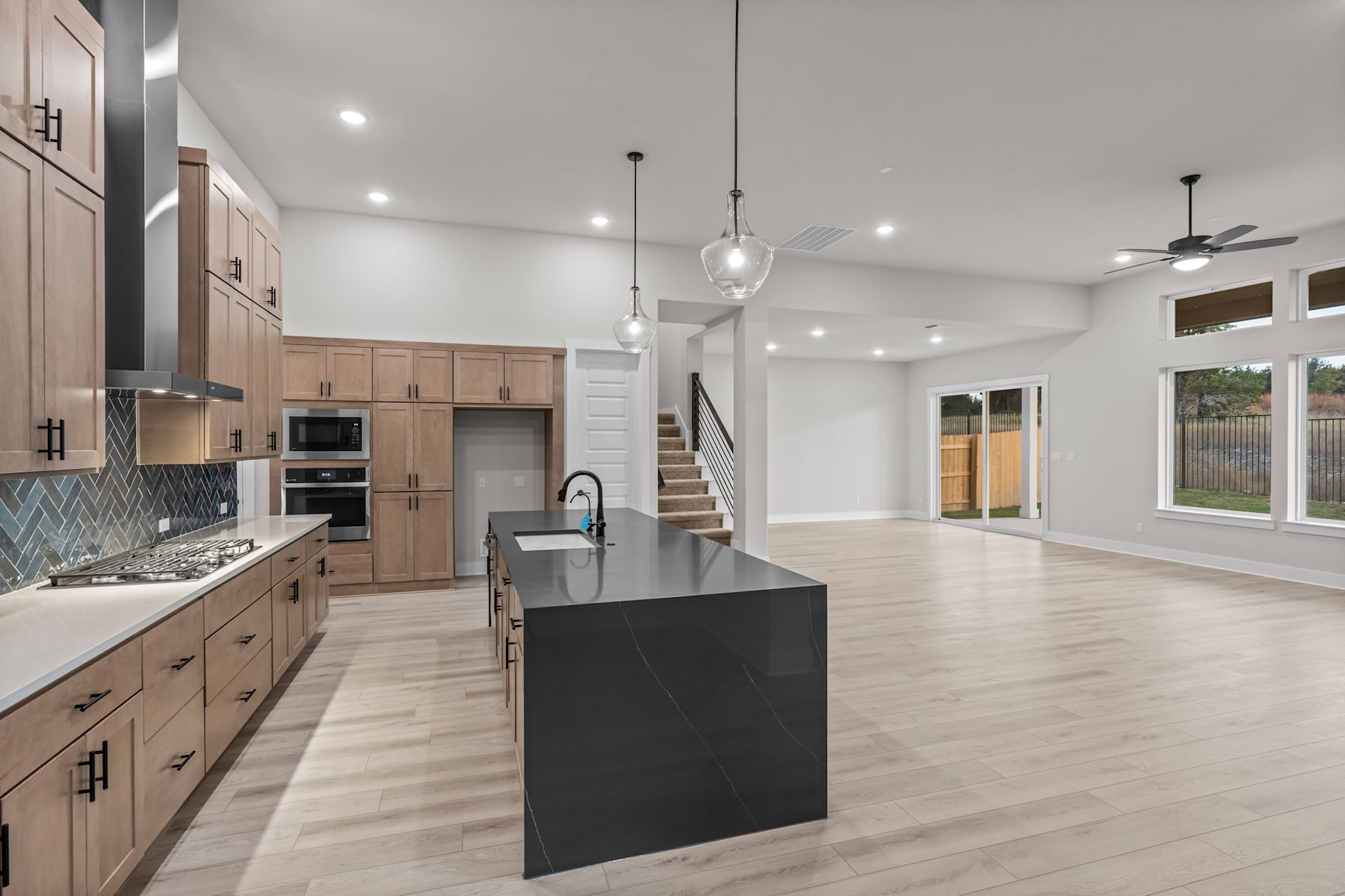 A modern, open-concept kitchen with light wood cabinetry, a large black island, and a bright, airy living space with hardwood floors and large windows.
