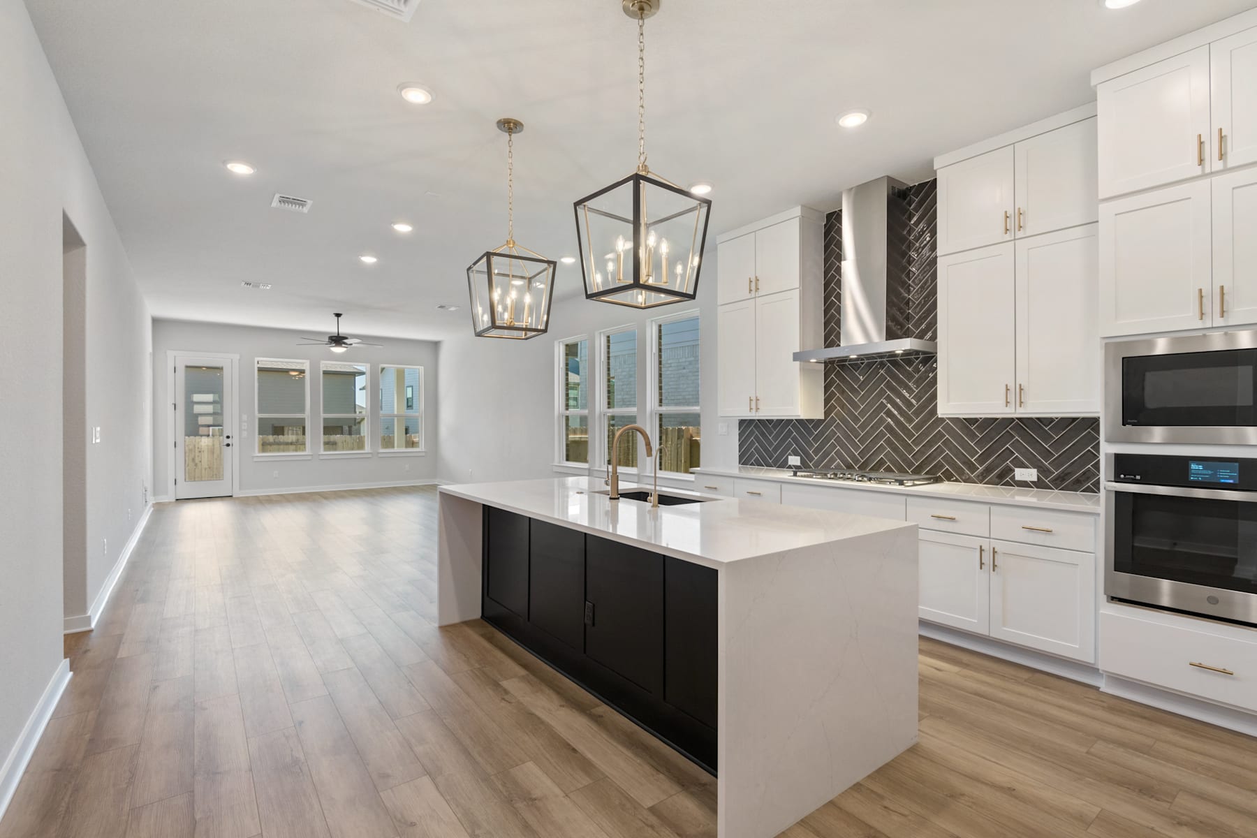 A modern, open-concept kitchen with white cabinets, a black island, and pendant lighting fixtures, set against a hardwood floor and leading into an adjacent room.