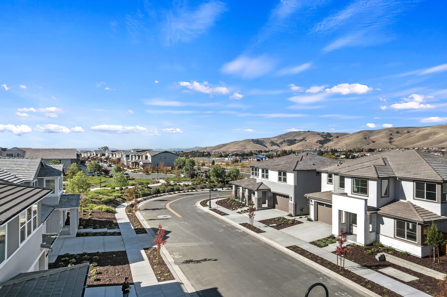 A residential neighborhood nestled in a hilly landscape, with rows of houses, winding roads, and a backdrop of mountains under a bright blue sky with scattered clouds.