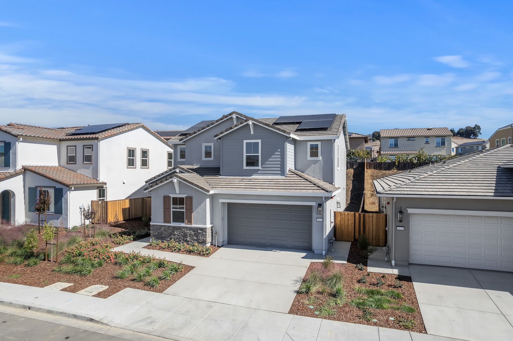 The image depicts a residential neighborhood with several modern, two-story houses featuring gray siding, gabled roofs, and attached garages. The foreground shows a well-maintained yard with landscaping, including small shrubs and plants. The background features a clear blue sky with scattered clouds, creating a pleasant and inviting atmosphere.