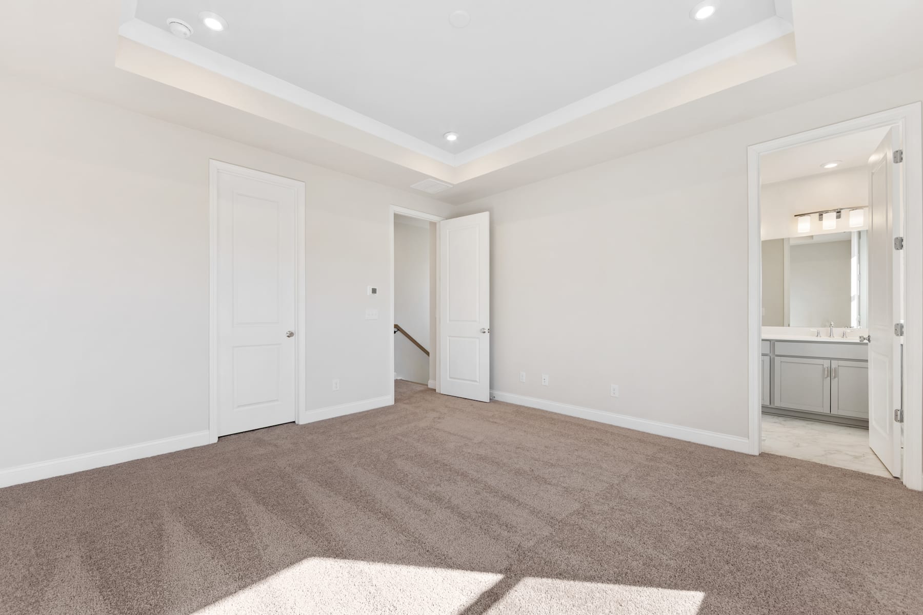 A spacious, well-lit room with white walls, a tray ceiling, and a carpeted floor. The room appears to be a bedroom or living space, with a doorway leading to another room visible in the background.