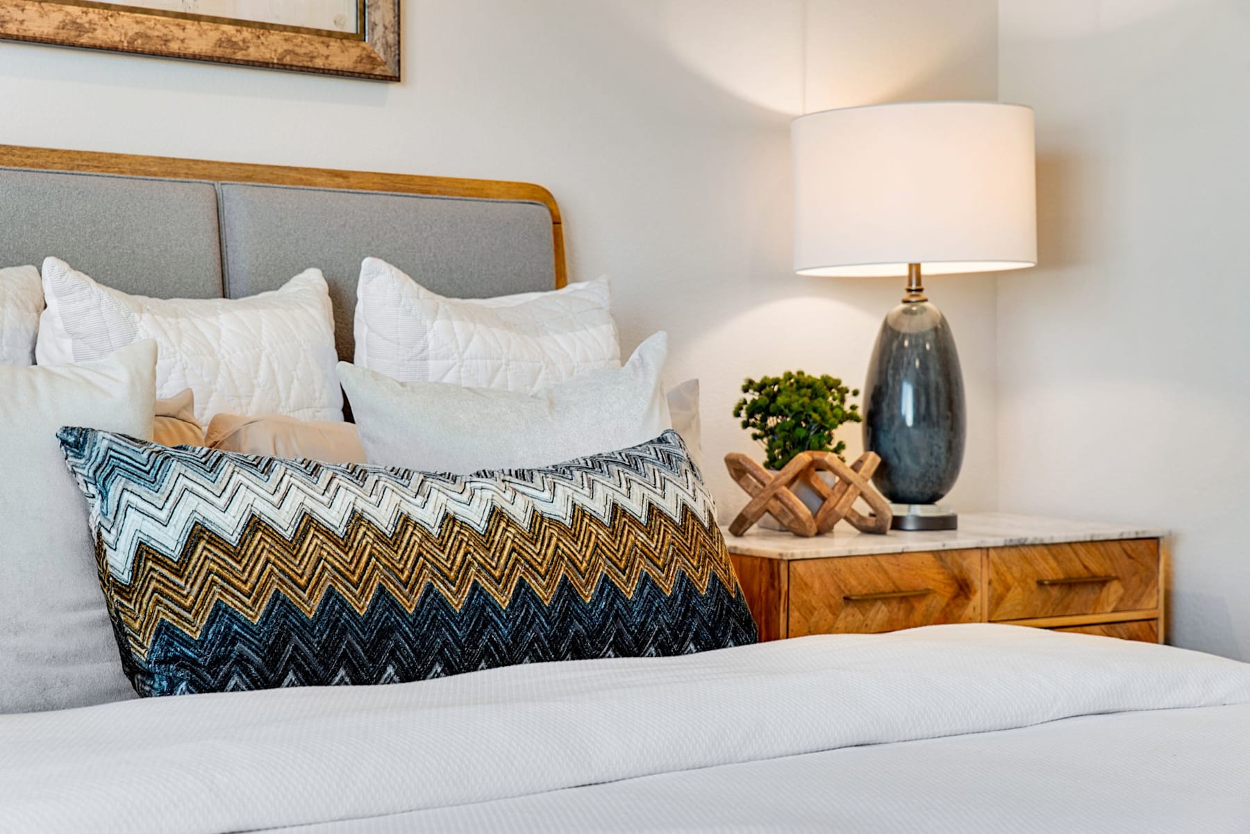 A cozy and inviting bedroom with a plush bed adorned with patterned pillows, a wooden nightstand with a lamp, and a framed artwork on the wall, creating a warm and stylish atmosphere.