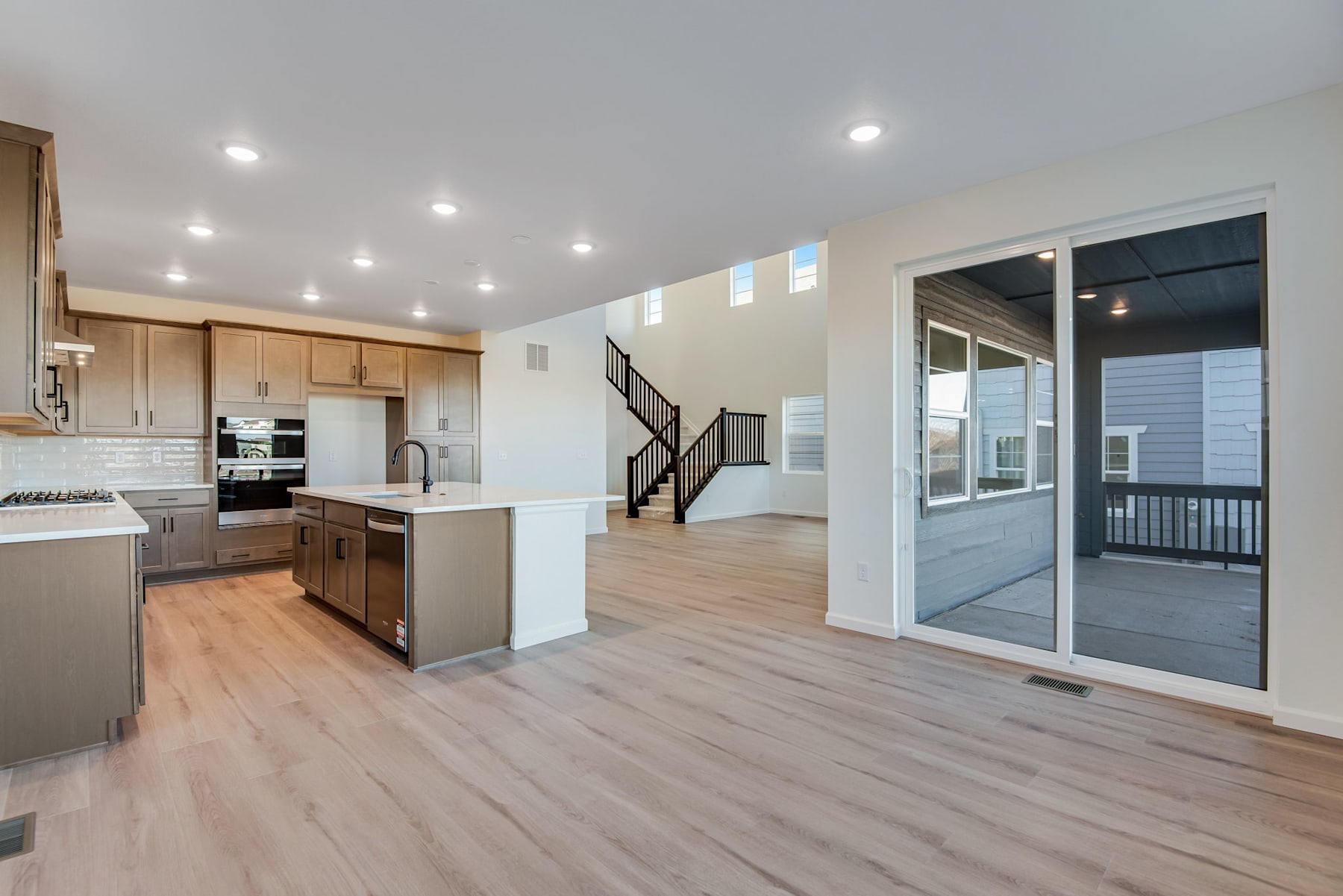 A modern, open-concept kitchen and living room with light-colored wood floors, white walls, and recessed lighting. The kitchen features light-colored cabinets and stainless steel appliances, while the living room has a sliding glass door leading to a balcony or patio.