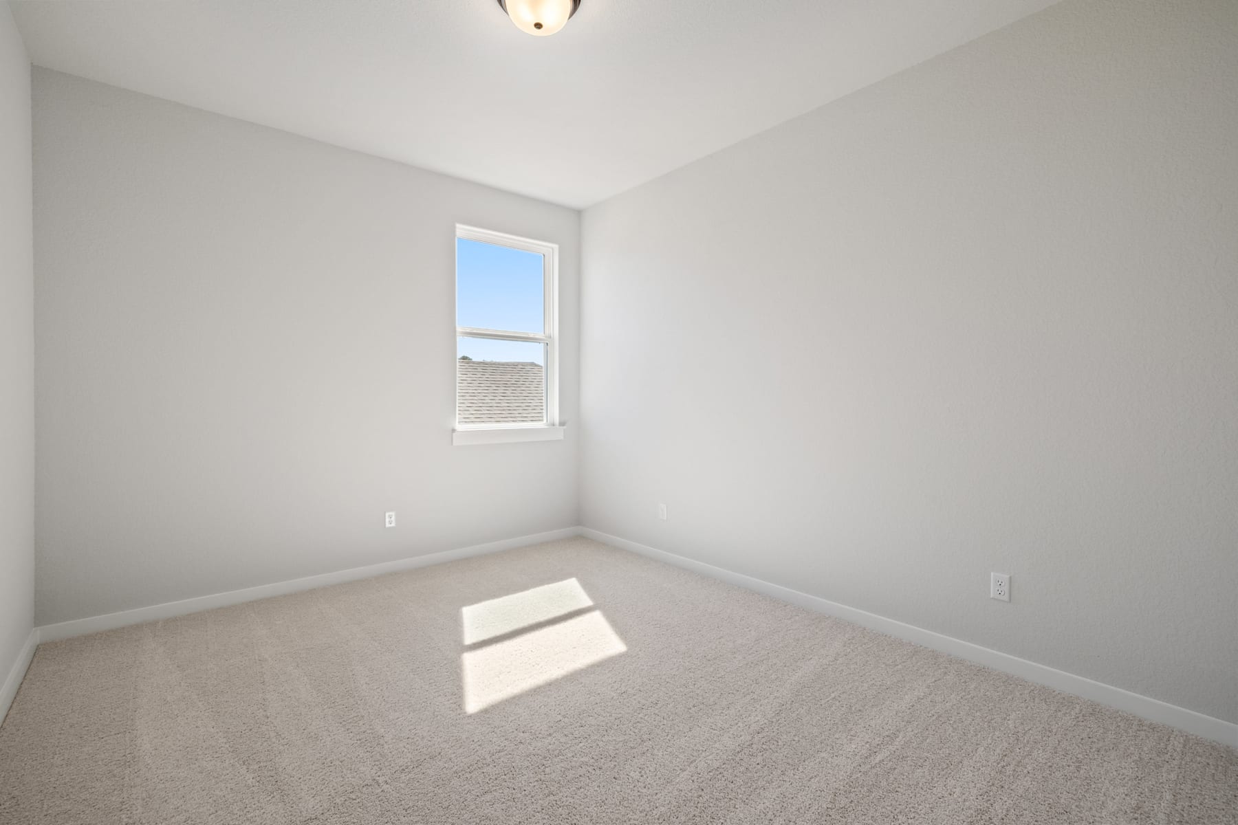 A bright, empty room with a window and a light fixture, featuring a plain white wall and gray carpeted floor.