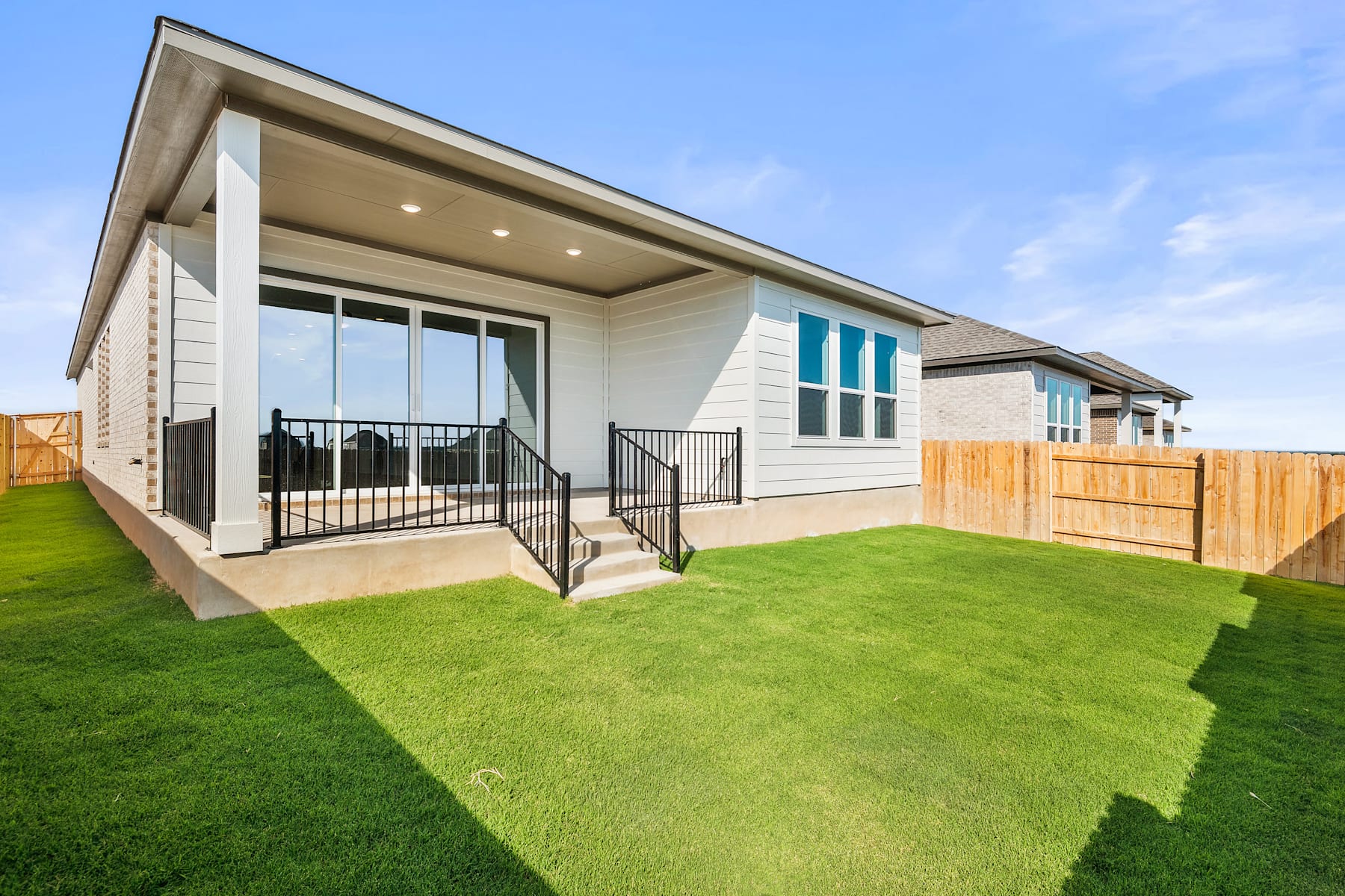 A modern, single-story house with a large, well-manicured lawn in the foreground, surrounded by a wooden fence and set against a clear blue sky.