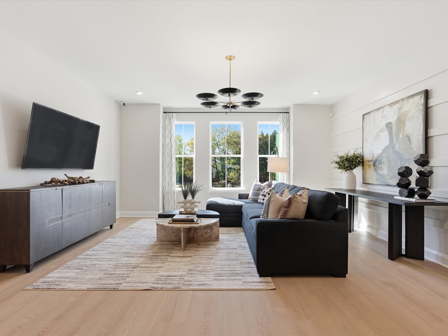 A modern and spacious living room with a large window, a dark sofa, a wooden coffee table, and a decorative chandelier overhead.