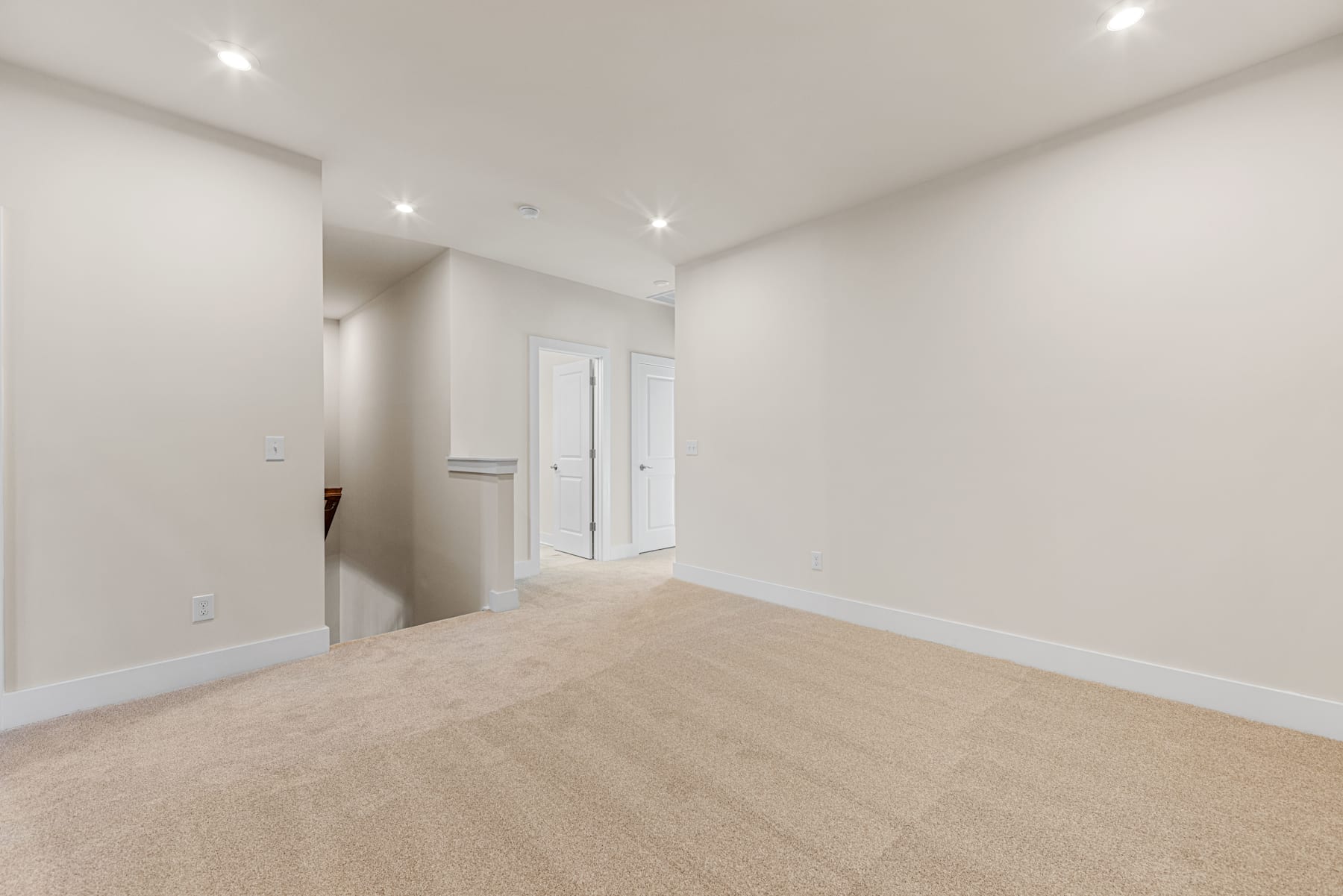 A spacious, well-lit room with a beige carpeted floor and white walls, featuring a doorway leading to another area.