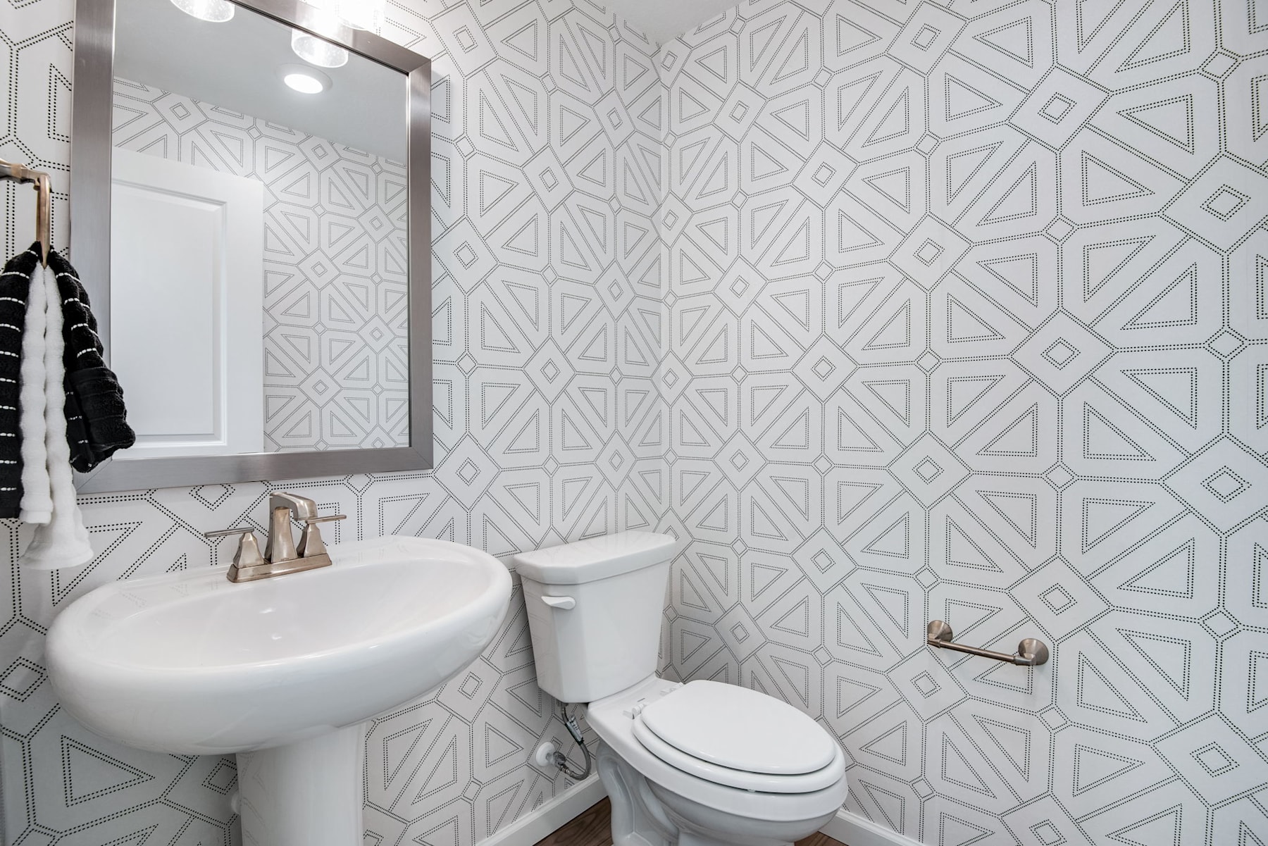 A modern bathroom with a white pedestal sink, a toilet, and a patterned wallpaper featuring geometric shapes in shades of gray.