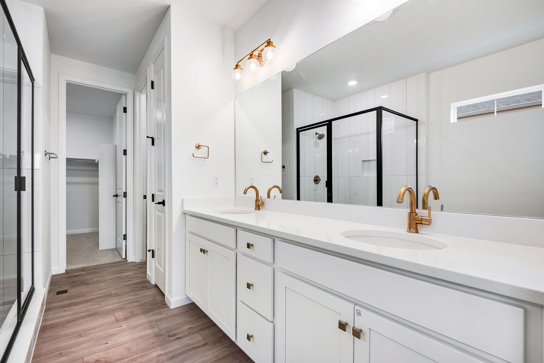 A modern, well-lit bathroom with white cabinets, a double vanity, and gold-toned fixtures, set against a backdrop of white walls and a wooden floor.