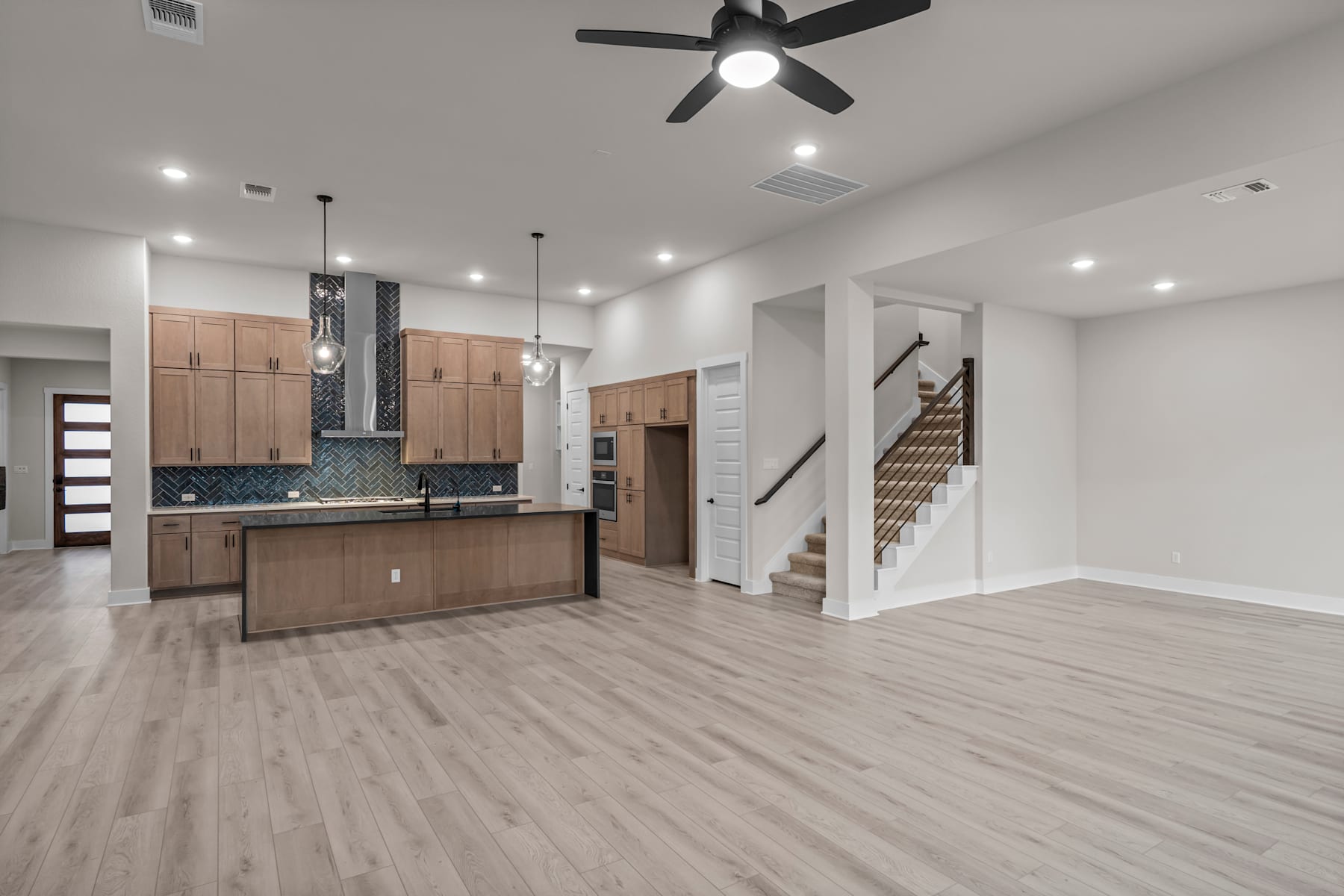 A modern, open-concept kitchen and living space with light-colored wood floors, wooden cabinets, and a ceiling fan.