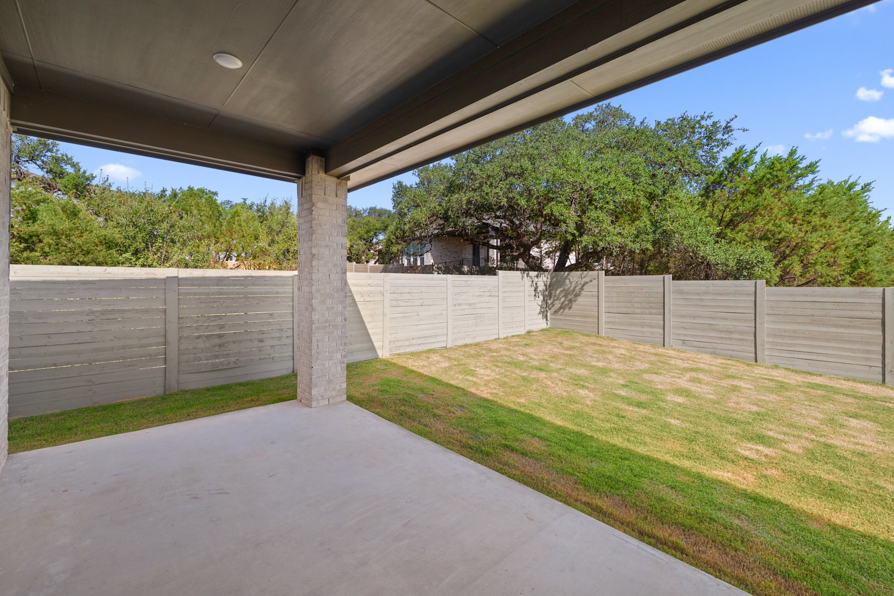 A covered patio with a grassy yard and a wooden fence surrounding the property, with trees and greenery visible in the background.