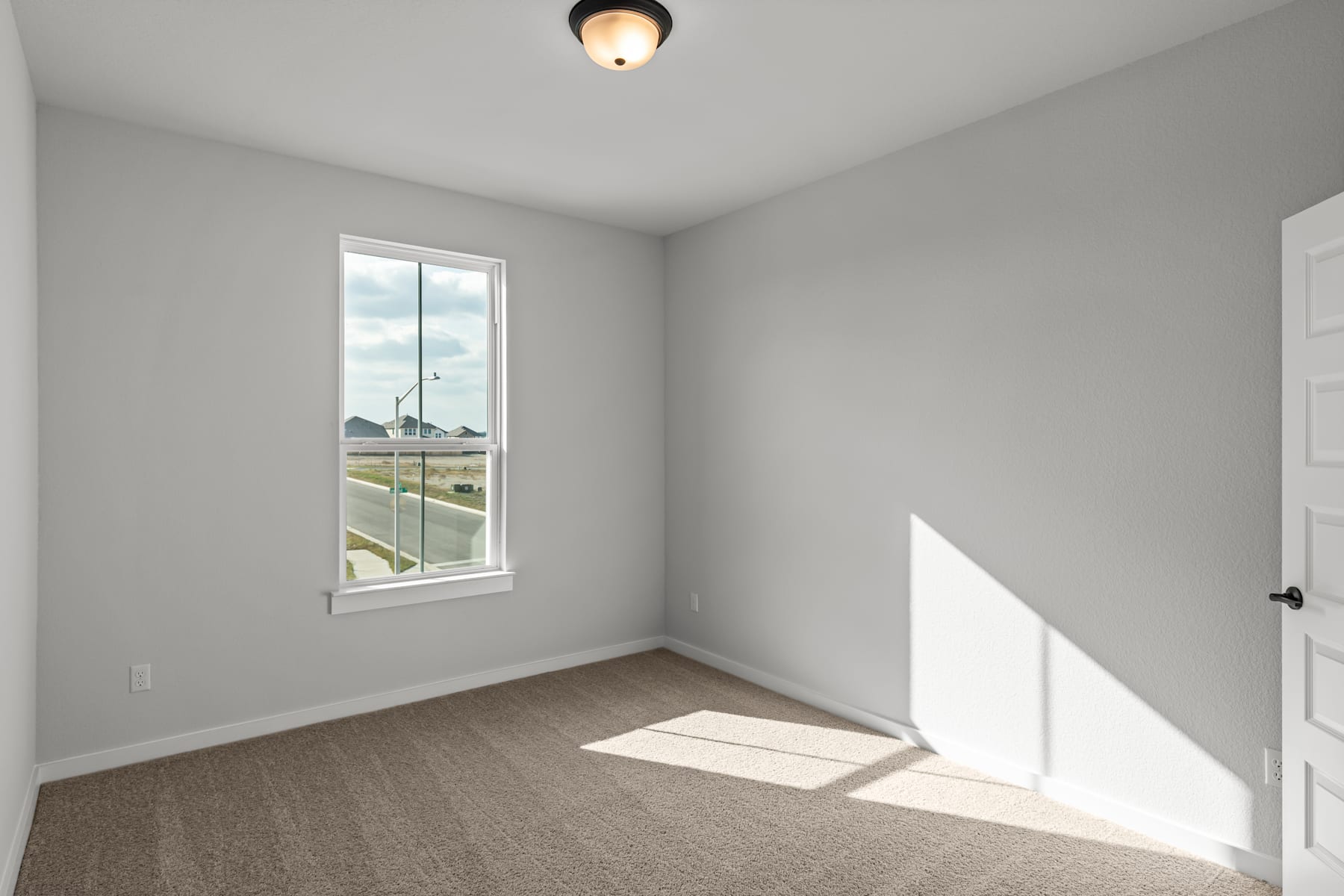 A sparsely furnished room with a window overlooking a grassy outdoor area, featuring a ceiling light fixture and a carpeted floor.
