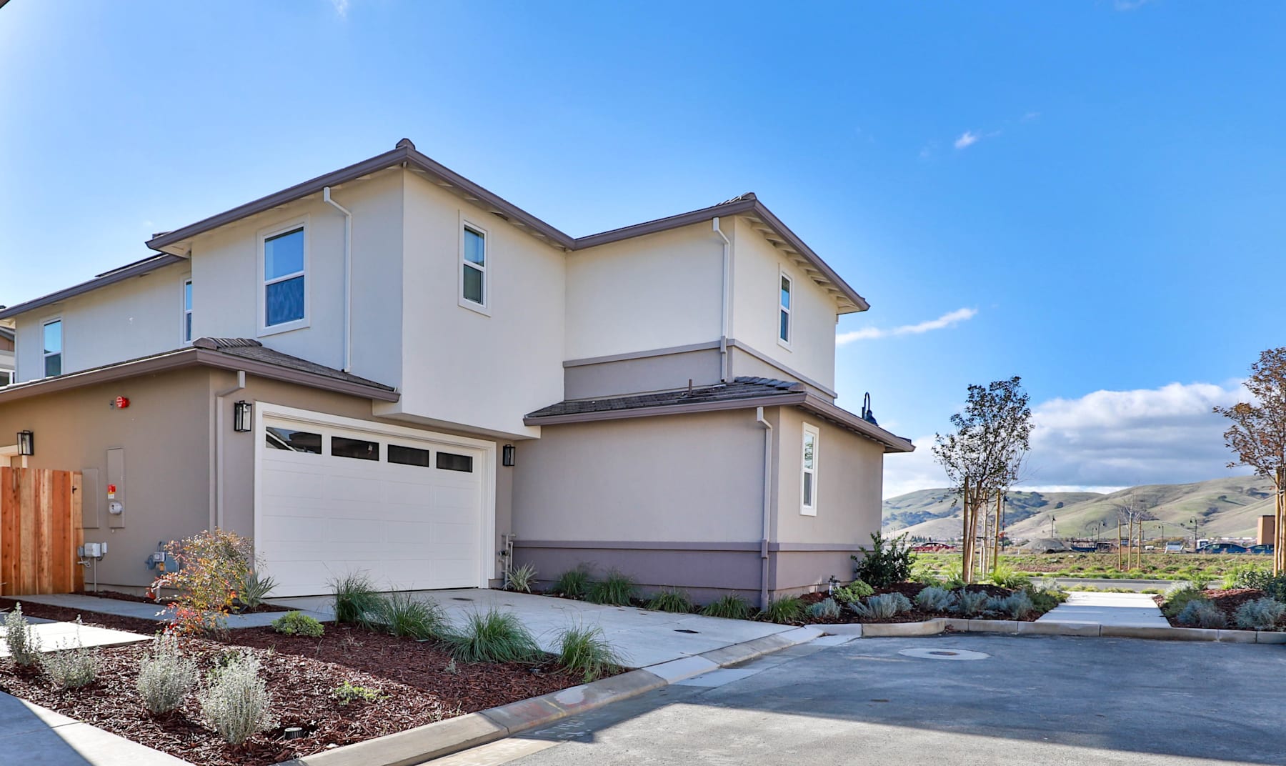 A modern two-story residential house with a garage door, surrounded by landscaped gardens and a paved driveway, set against a clear blue sky with some clouds.