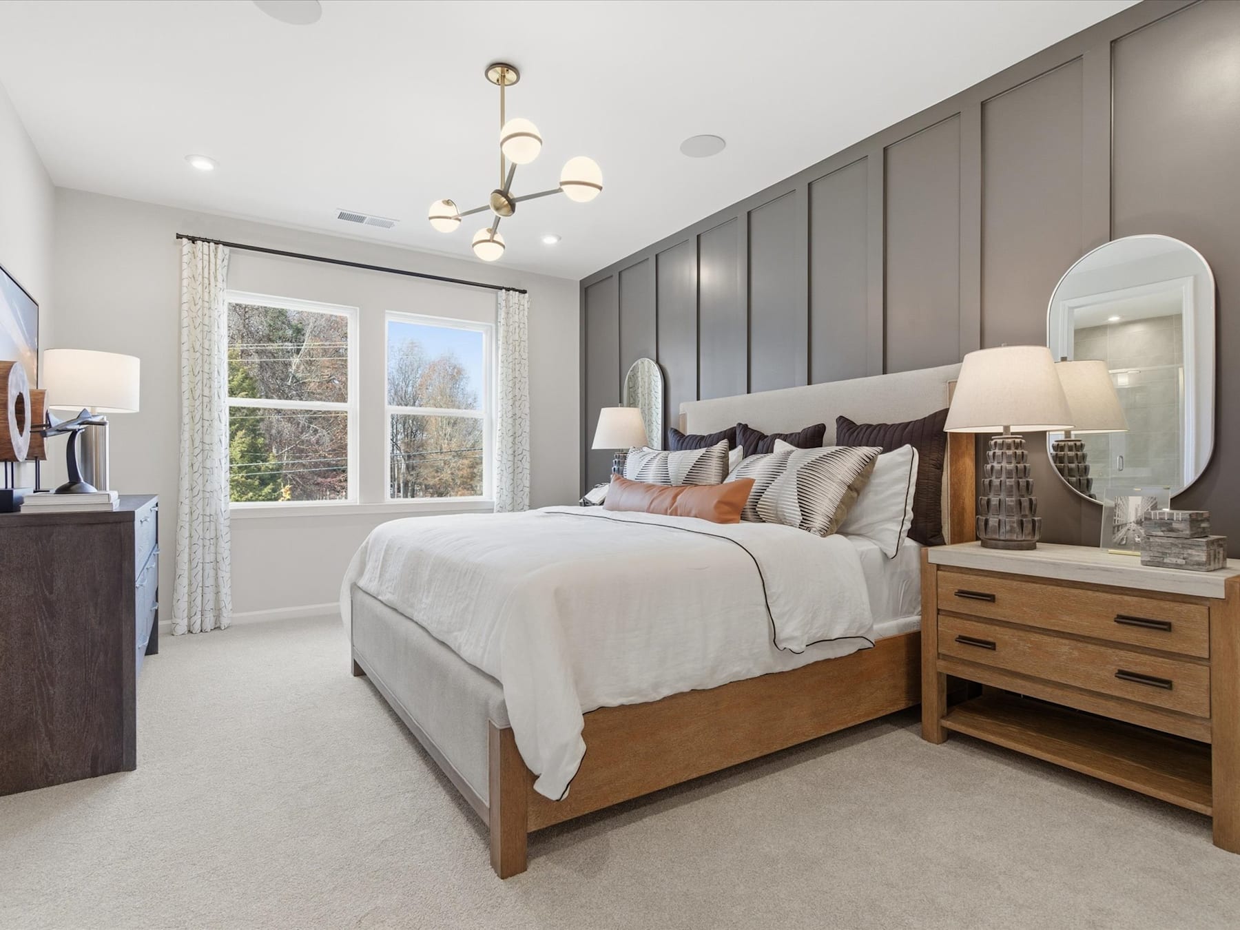 A cozy and well-appointed bedroom with a large bed, wooden furniture, and a chandelier overhead, set against a backdrop of large windows and a neutral color scheme.