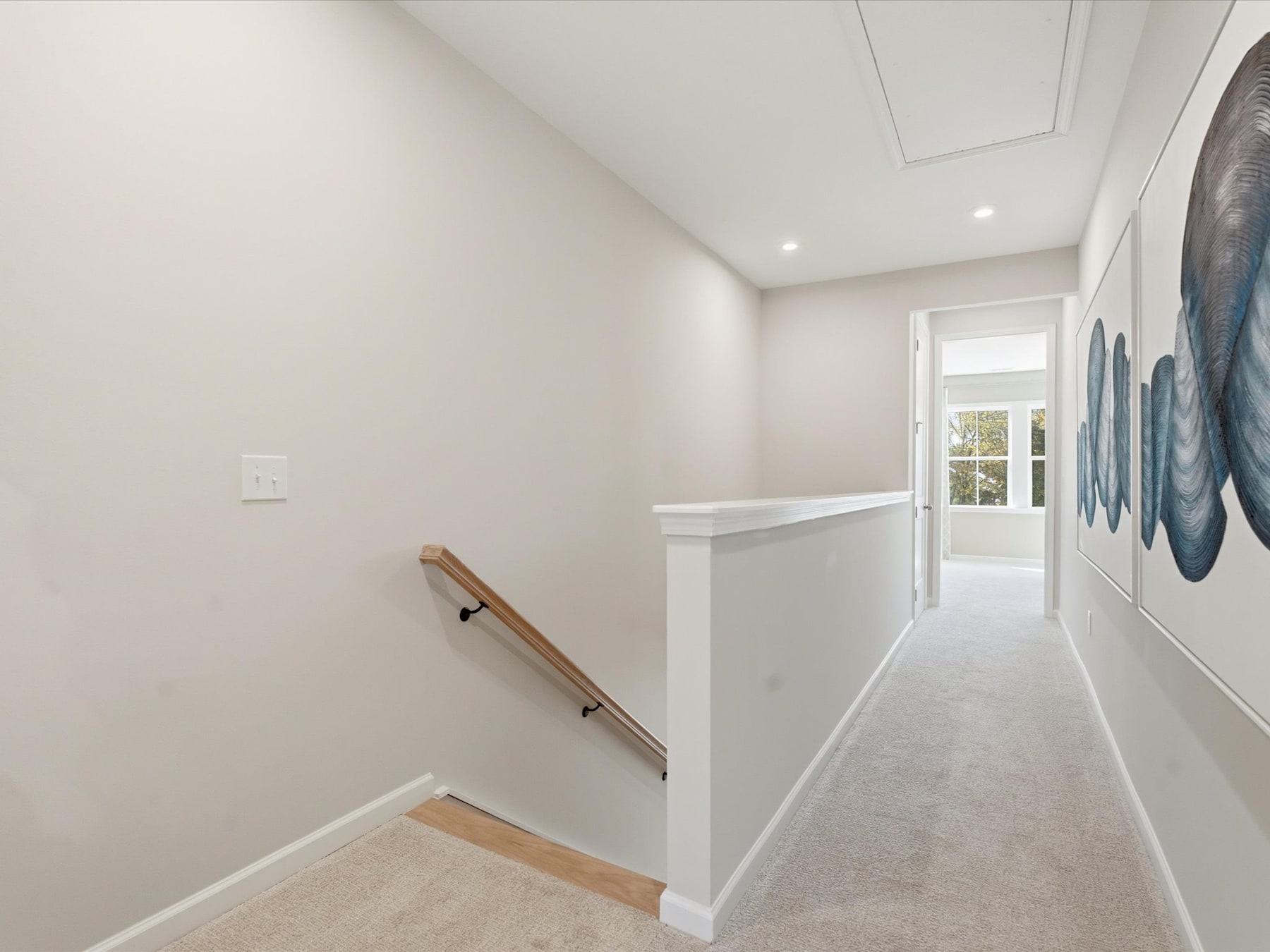 A bright, minimalist hallway with a wooden handrail leading to a doorway, and abstract artwork adorning the walls.