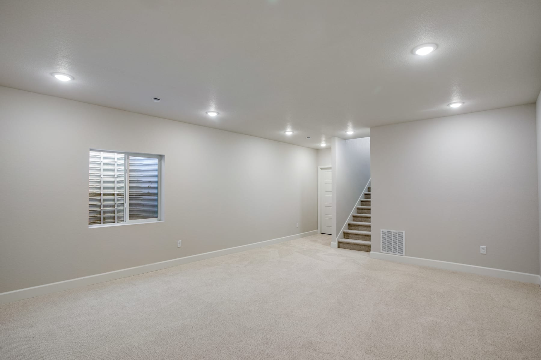 A spacious, well-lit room with a staircase leading to an upper level, featuring a large window and recessed lighting in the ceiling.
