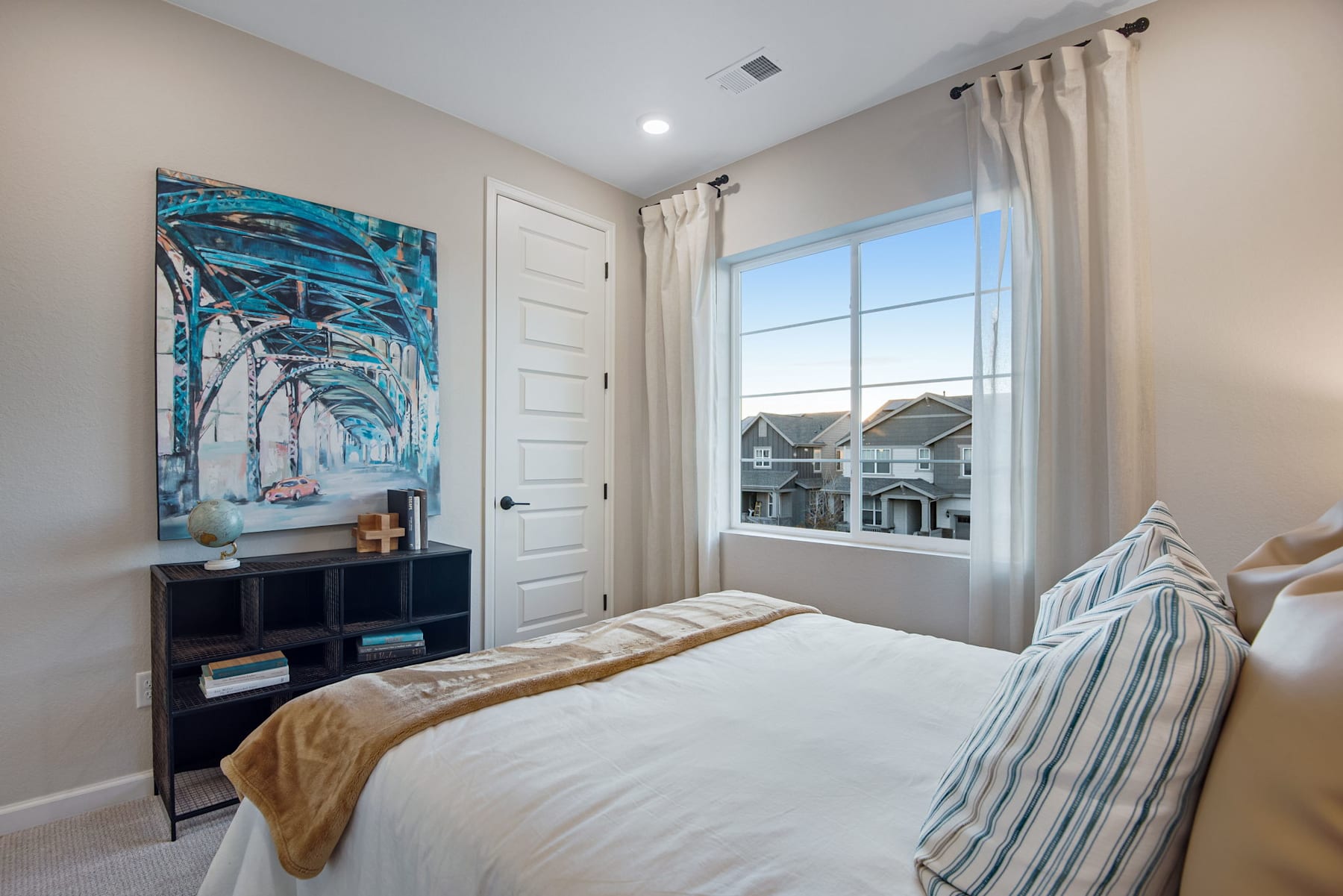A cozy and well-furnished bedroom with a large window overlooking a residential neighborhood, featuring a vibrant abstract painting on the wall and a comfortable-looking bed with striped bedding.