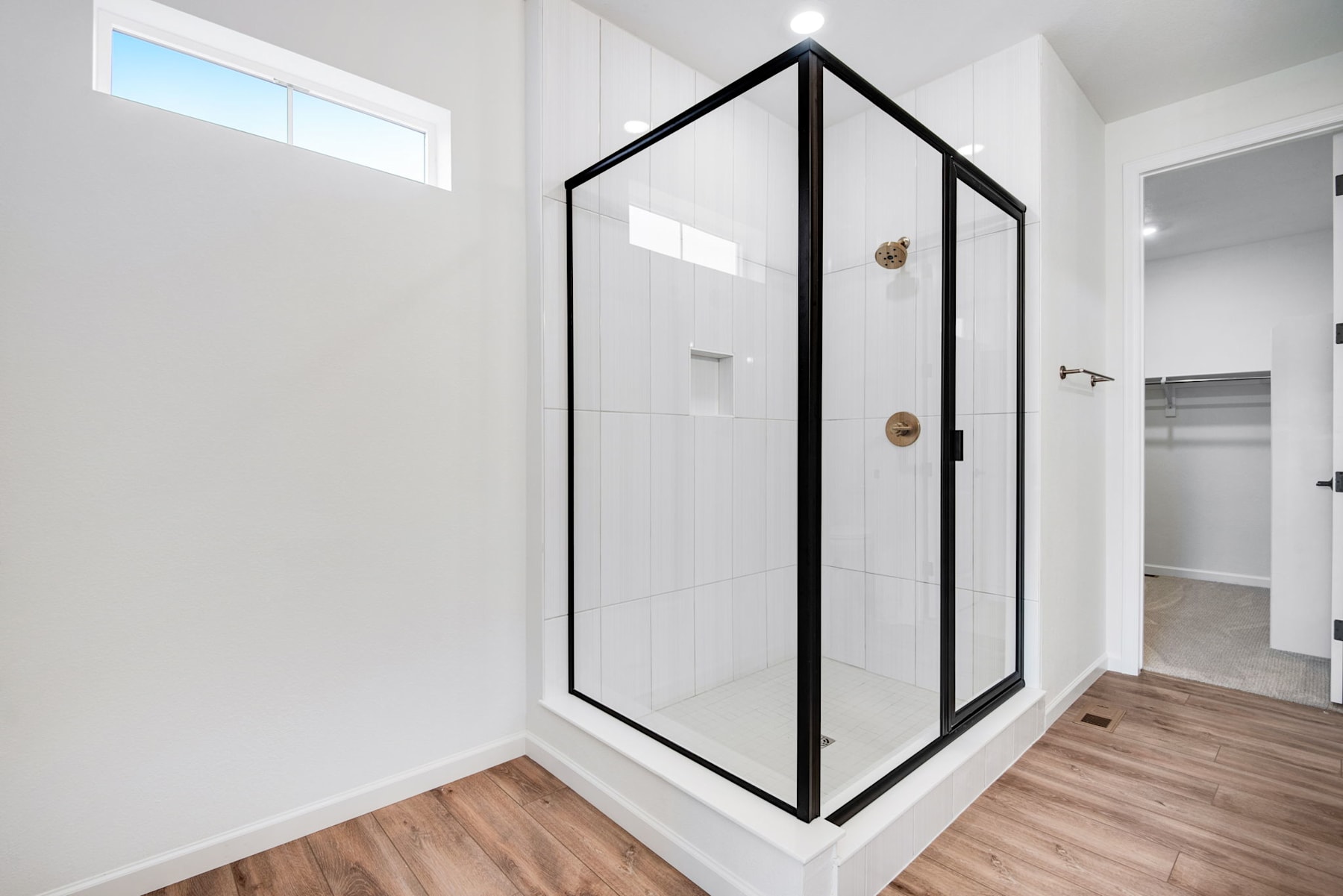 A modern, minimalist bathroom with a glass shower enclosure framed in black metal, set against a bright, white-walled background and wooden flooring.