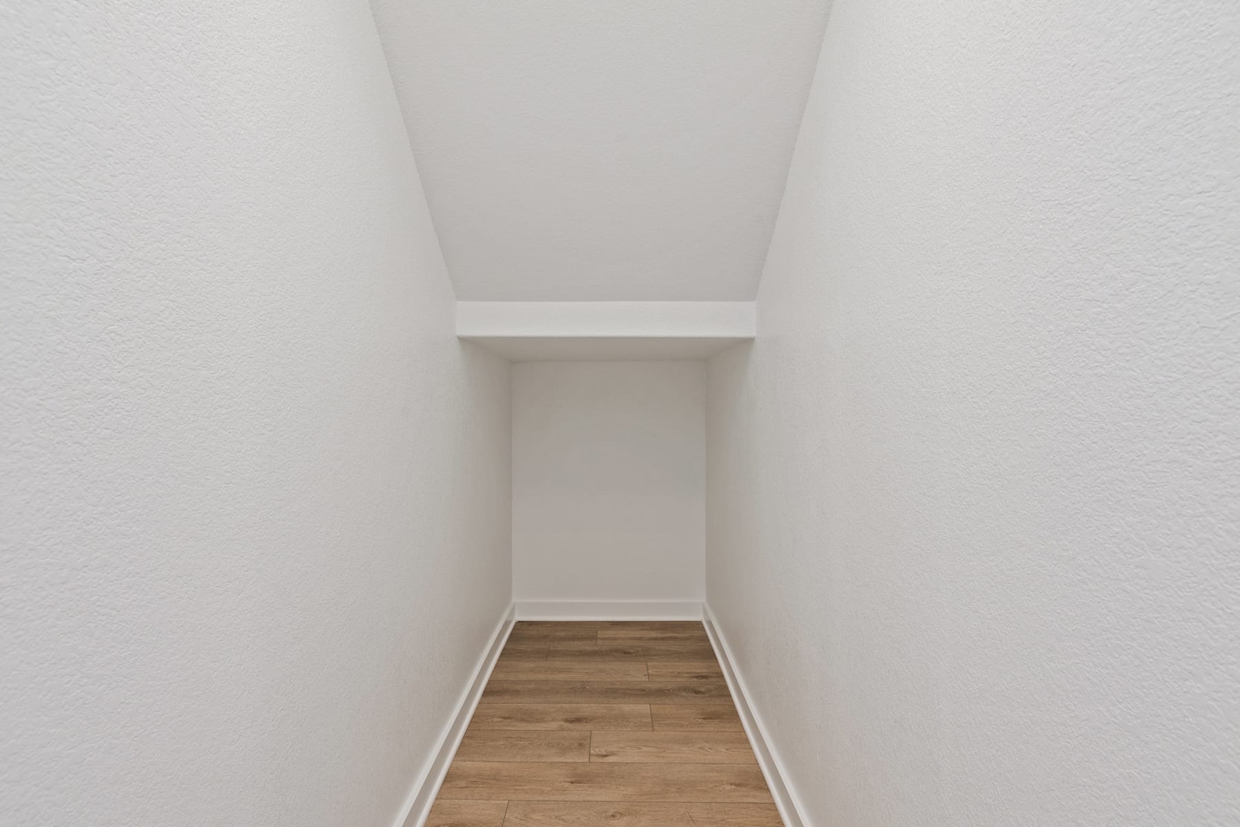A narrow, well-lit hallway with a wooden floor and white walls leading to an open doorway.