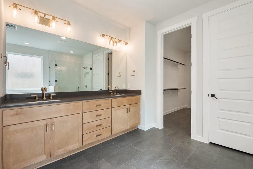A spacious bathroom with a large vanity, dark countertop, and recessed lighting fixtures, leading to an open doorway in the background.