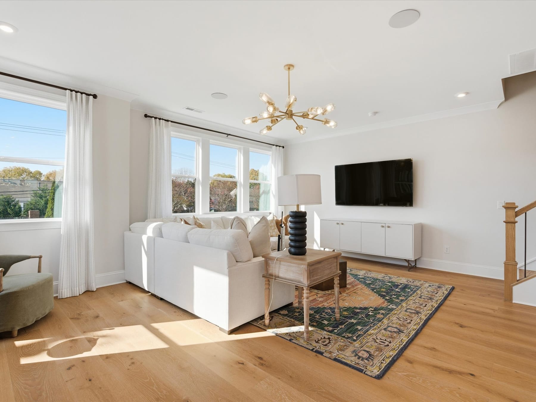 A bright and airy living room with white furniture, a large window, and a chandelier hanging from the ceiling. The room features a patterned rug and a television mounted on the wall.